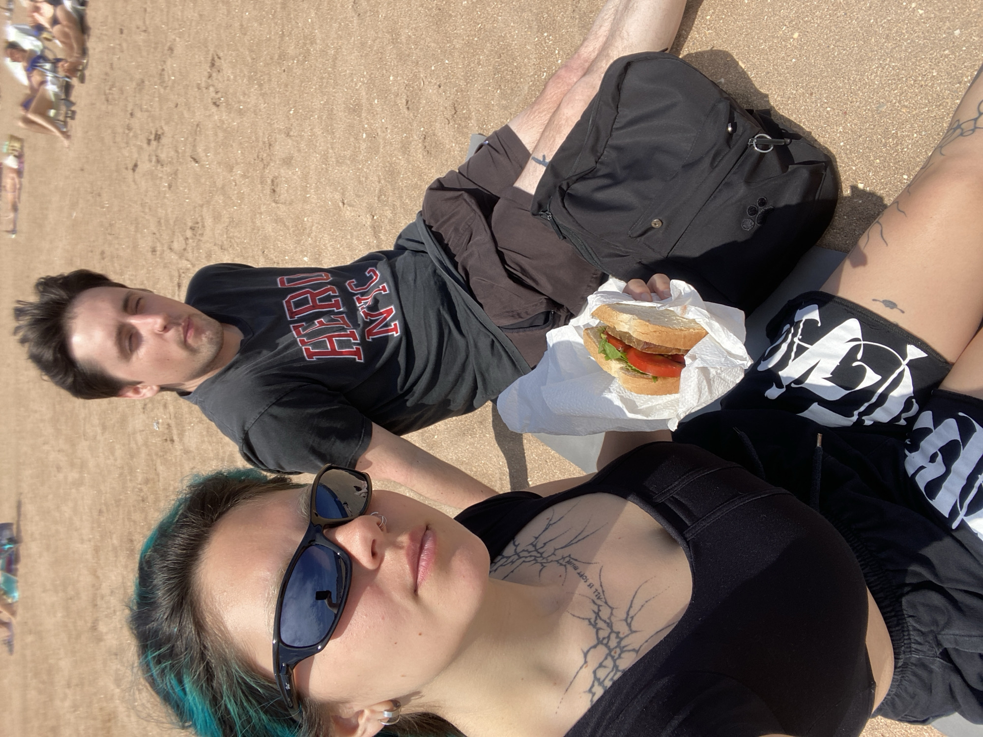 Two people are lying on a sandy beach, likely in Gijón/Xixón, Spain, given the light-colored sand characteristic of the region. A woman with blue-tinted hair and sunglasses is in the foreground. She has tattoos on her neck and upper chest, including script. Behind her, a man is lying down, wearing a black t-shirt with "HERO NYC" in red and white lettering. He is looking up with closed eyes. Adjacent to him is a sandwich wrapped in white paper. To the left of the frame, other people are visible on the beach. The sand appears dry. A backpack is also present.