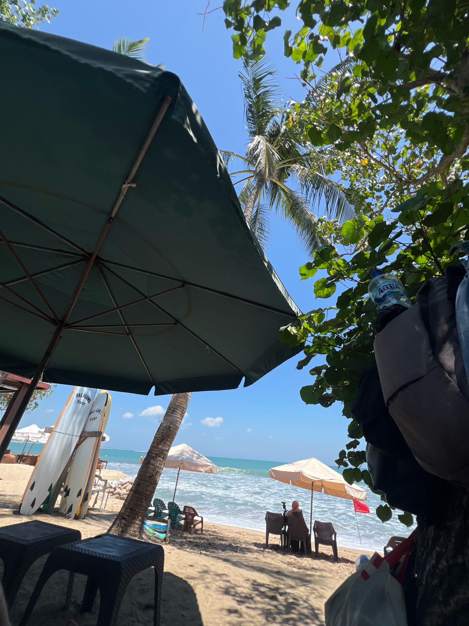 This is a low-angle, eye-level, full outdoor shot taken on a beach in Kuta, Indonesia, on a bright, sunny day. The foreground is dominated by a large, dark green beach umbrella, casting a shadow. To the left and slightly behind it are two black woven plastic tables. Leaning against a nearby structure are two surfboards, one white with orange accents and black text that reads "BALIGUNS SURF" and another yellow surfboard, also with the same text. Behind these, several white beach chairs are visible.
In the midground, the sandy beach stretches towards the turquoise ocean. A light beige umbrella stands open, with two chairs positioned beneath it, occupied by one person wearing a headscarf. Next to these chairs, a small red flag with white writing is planted in the sand. Further to the right, a palm tree trunk is prominent.
The background shows the ocean with moderate waves and a clear blue sky with scattered white clouds. Lush green foliage from trees hangs into the frame from the upper right, with some sunlight filtering through the leaves. A partially visible backpack with a water bottle in a side pocket is also present on the right side of the frame, attached to a tree trunk. The overall impression is of a relaxed beach scene with an emphasis on sun, sand, and water activities.