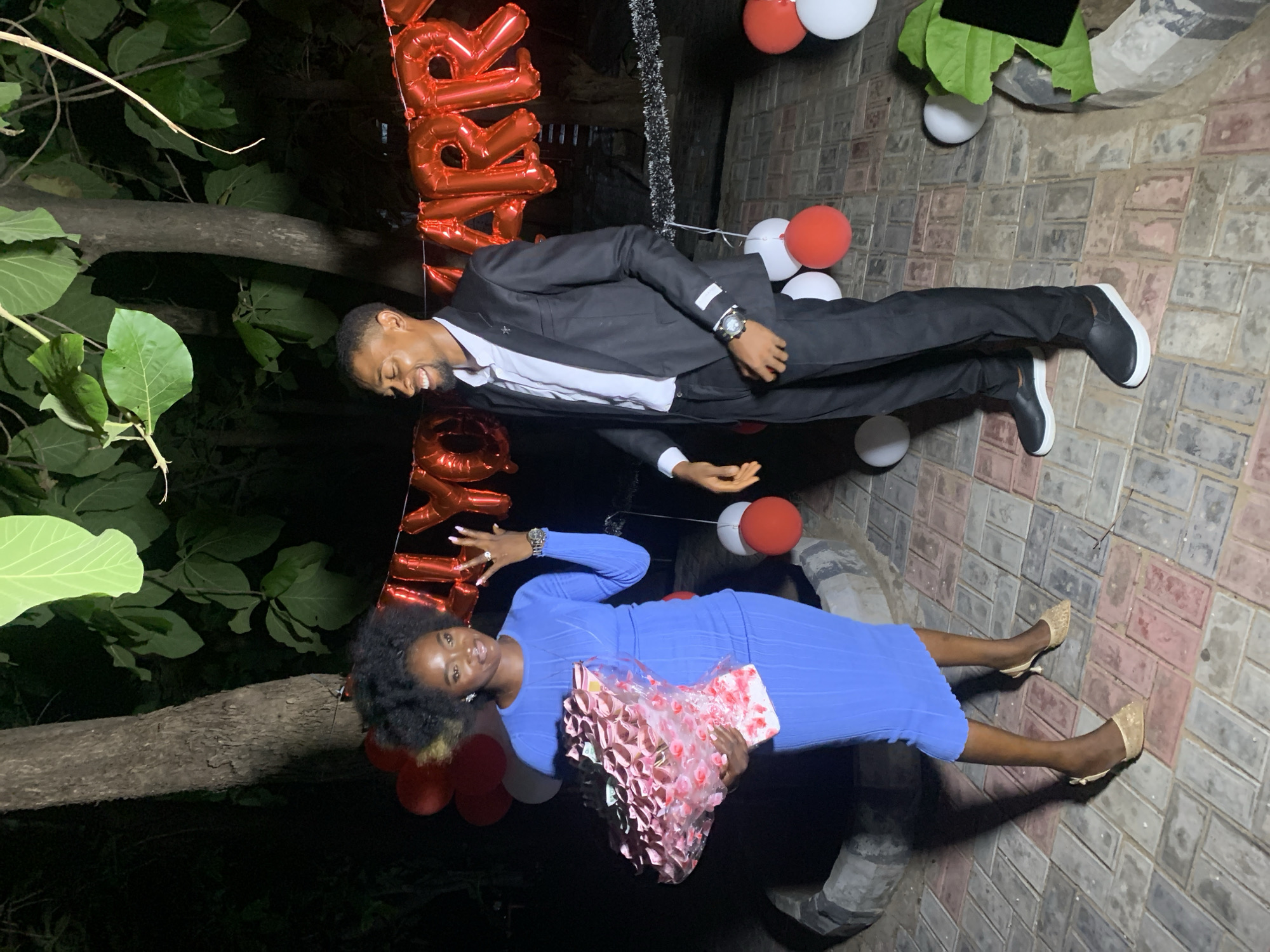 The image depicts a man and a woman standing next to each other outdoors in Adankolo, Nigeria. The woman is wearing a long-sleeved blue dress and heels, holding a wrapped bouquet. The man is wearing a black suit and white shirt. Both are standing on a brick-paved area and there are balloons in the background. Trees and foliage are visible in the surrounding area.