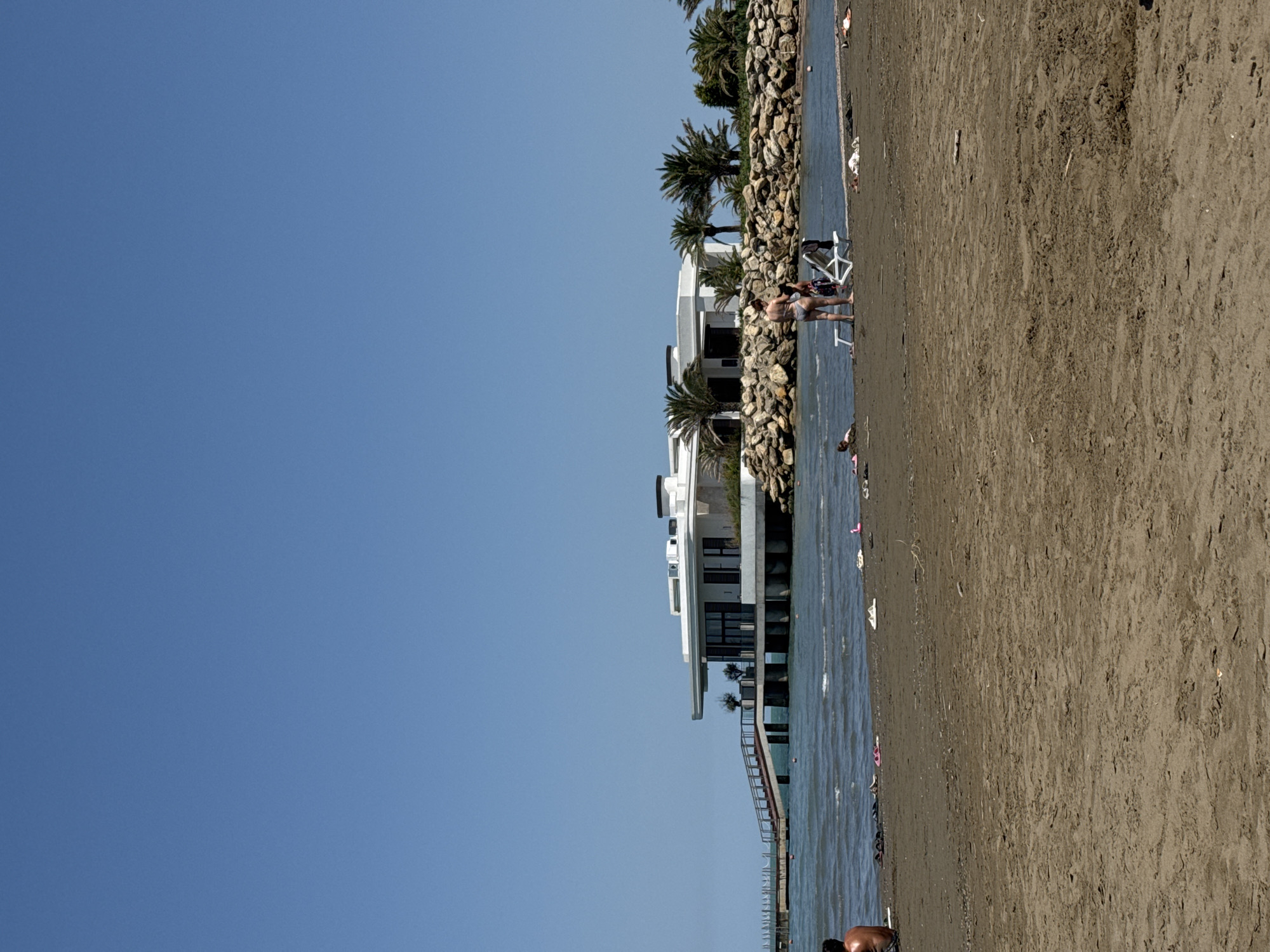 This is a medium-shot, eye-level, vertical, outdoor, daytime, photographic image of a beach scene in Sabunchu Raion, Azerbaijan, likely near a resort or restaurant. The foreground features a sandy beach with dark, wet sand. In the middle ground, the water of the Caspian Sea is visible, with small waves and some debris floating. Several people are scattered across the beach. To the left, a person with their back to the camera is seated on the sand. Further into the water, there are more people and some floating objects. In the mid-right of the image, two individuals are standing near beach chairs, one wearing a bikini and the other a patterned dress. In the background, a modern, white, multi-story building with large windows and a flat roof stands elevated on stilts over the water. A ramp with a railing leads up to the building. The building is surrounded by a rock barrier, with palm trees and other vegetation visible behind it. The sky is clear and blue, with no clouds. A pier extends into the water on the far left of the image. The overall scene depicts a sunny day at a beach resort.