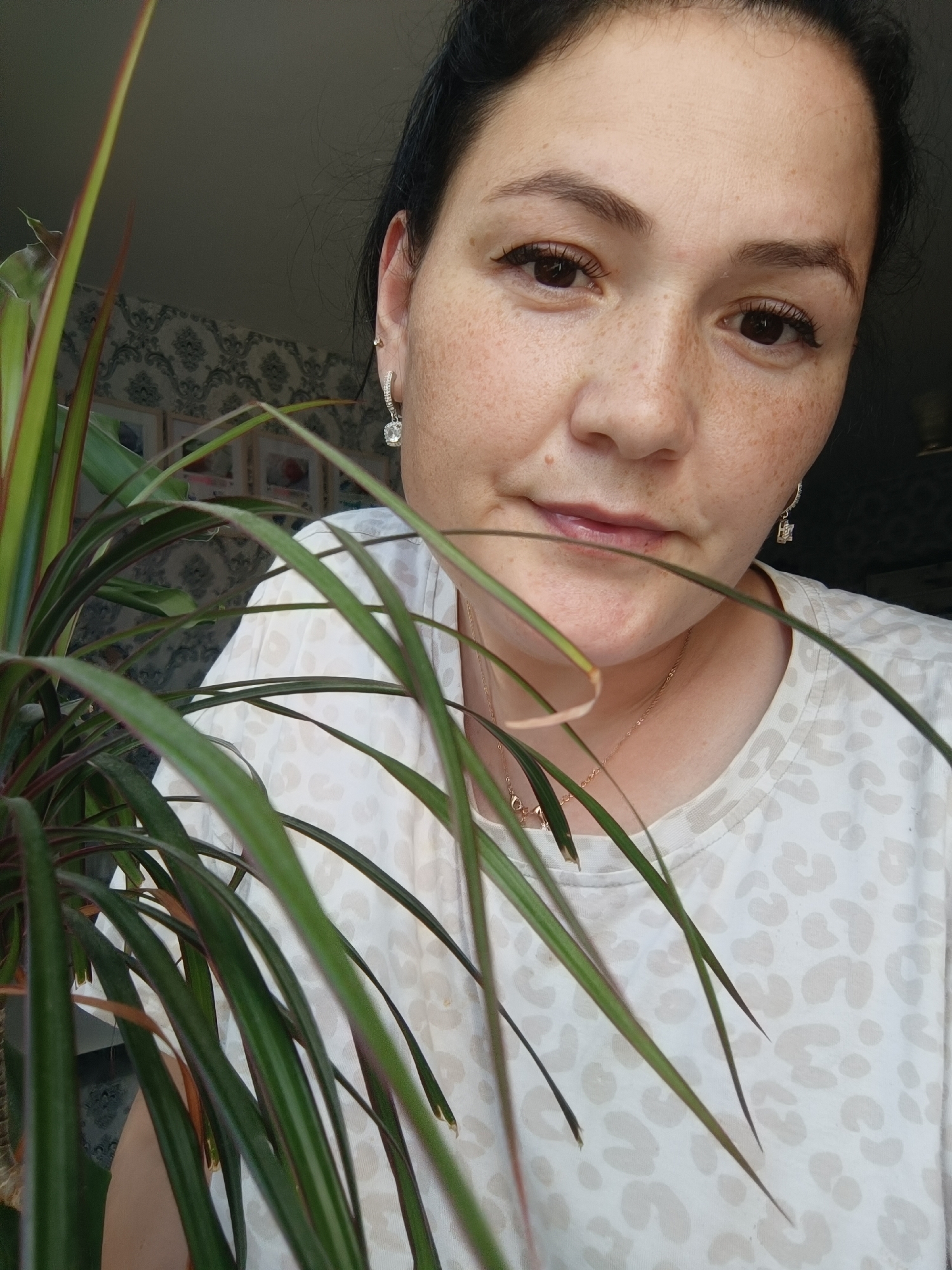 A woman with dark hair and freckles is looking at the camera from behind a leafy plant. She is wearing a white t-shirt with a leopard print pattern and a necklace. She has earrings in her ears. The background shows a patterned wallpaper and framed pictures. The image appears to be taken indoors, possibly in a home in Chaikovsky, Russia.