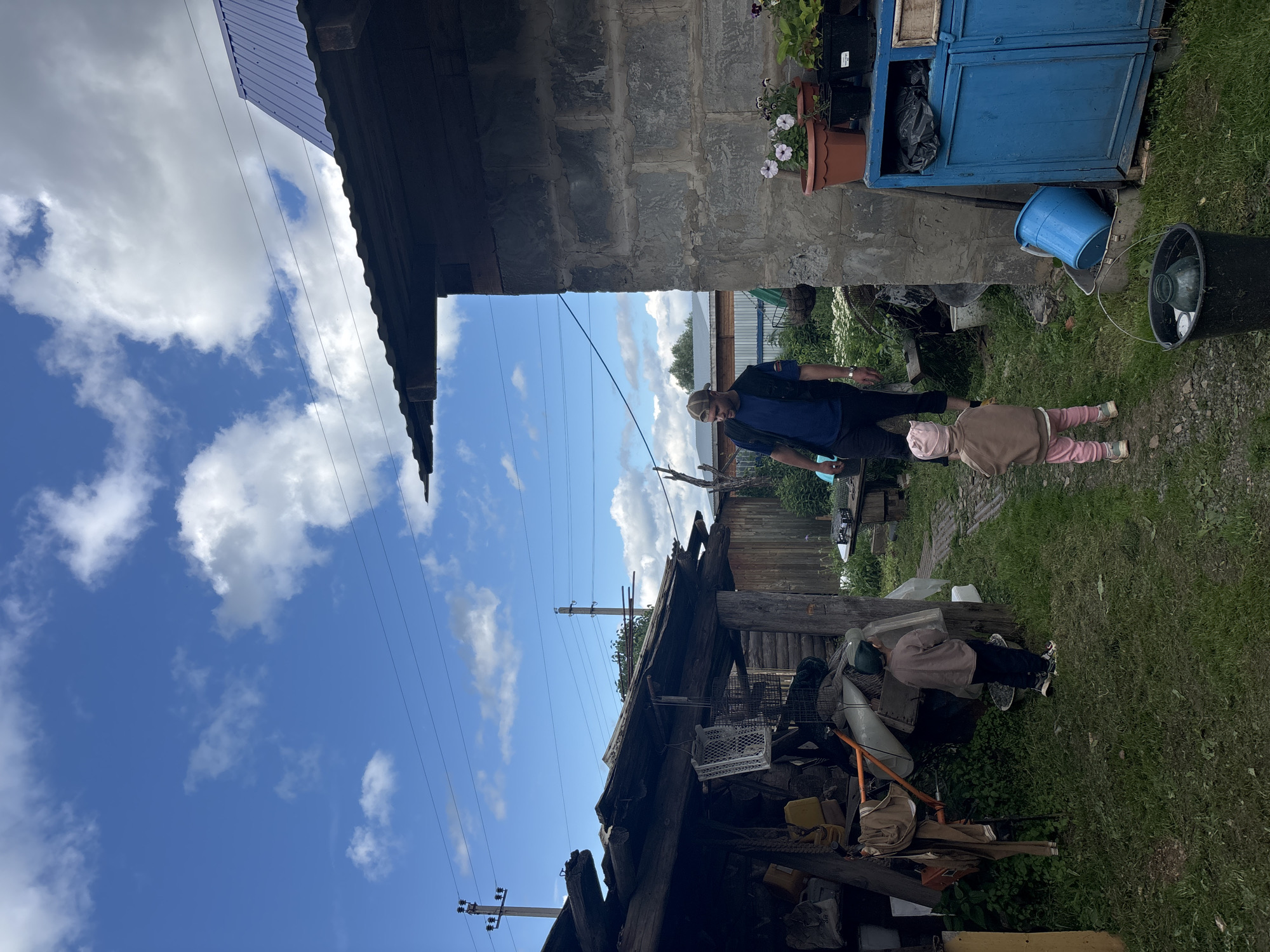 This image depicts an outdoor scene, likely in a rural setting in Niva, Russia, judging by the architecture and general atmosphere. The day appears to be sunny with scattered clouds, suggesting fair weather.
The primary subjects are two individuals: an adult and a young child. The adult, wearing a dark jacket and a cap, is standing and appears to be interacting with the child. The child, dressed in a light pink hat and outfit, is lying on the ground facing away from the camera. Their posture and clothing suggest a casual or playful moment.
The scene is characterized by a somewhat cluttered yard. Various objects are visible, including a blue cabinet, several buckets, and a pile of what appears to be discarded materials or old structures. A rustic wooden building with a damaged roof is prominent in the foreground, hinting at a possibly rundown or developing property. Power lines are visible crisscrossing the sky, a common sight in rural areas. In the background, there's a fence and some greenery, indicating a lived-in space.
There are no visible text elements within the image. The overall impression is one of everyday life in a rural environment, with the presence of both people and the accumulation of objects typical of a household yard. The lighting suggests it is daytime, likely afternoon given the brightness.