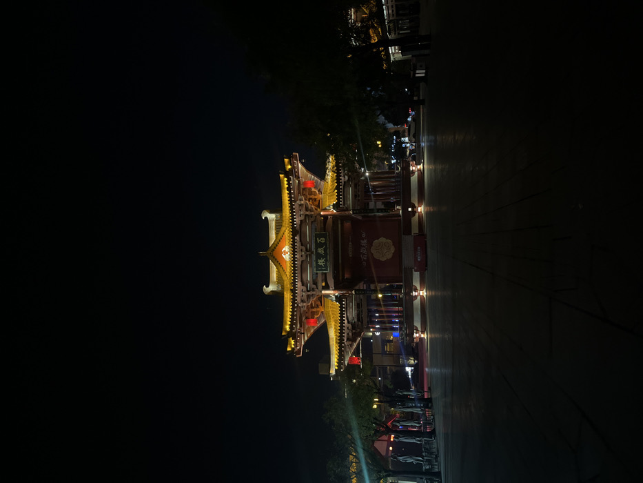 The image displays the Baisheng Lou (百盛樓) pavilion situated within the Datang Everbright City in Yanta District, Xi'an, China, during nighttime hours. The central subject is a multi-tiered, traditional Chinese-style structure with prominently illuminated yellow and gold upturned eaves and multiple red hanging lanterns. A vertical sign bearing the Chinese characters "百盛樓" is centered on its facade. Below this sign, a large red panel or banner features a circular design with additional Chinese text. The structure is supported by dark columns and rests on a raised, illuminated base. In the foreground, a large, dark paved square extends towards the viewer, reflecting artificial light directly in front of the pavilion. To the left, indistinct outlines of outdoor seating, including tables, chairs, and folded umbrellas, are present under minimal illumination. Dark trees with some illuminated foliage flank both sides of the square. Distant urban structures with faint lights are visible in the background. No individuals or specific actions are discernible within the frame.