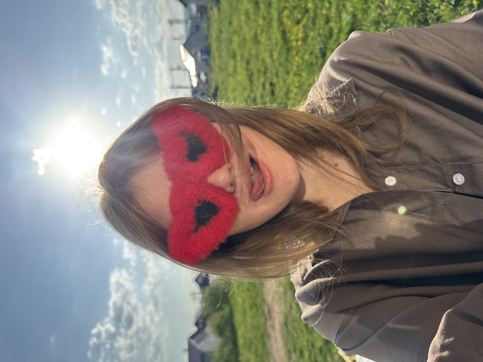 A young woman is outdoors in Ферма, Russia, wearing a furry, red butterfly-shaped eye mask. She is sticking her tongue out playfully. She is wearing a brown shirt with white buttons. The background includes a blue sky with clouds, green grass, and buildings in the distance. The sun is shining brightly behind her head.