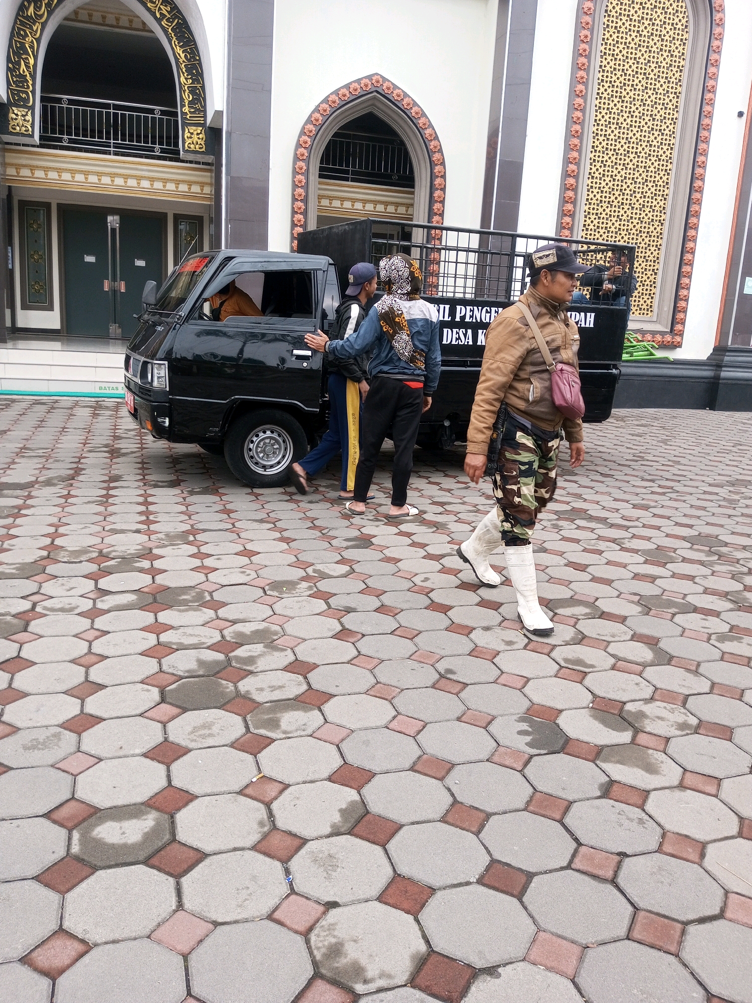 In the city of Keseneng, Indonesia, in front of a building with Islamic architectural elements, a black pickup truck is parked on an octagonal tiled ground. The building has white and grey walls, with arched entrances decorated with gold Islamic calligraphy and floral motifs in red and orange. The tiled ground is wet, indicating recent rain.
There are several individuals in the scene. A man in a brown jacket, camouflage pants, and white boots is walking away from the truck. He has a pink pouch slung across his shoulder. Another man is sitting in the driver's seat of the truck, wearing an orange shirt. Two other individuals, one in a blue jacket and the other in a black jacket with a patterned scarf, are standing near the open driver's side door of the truck. The individual in the blue jacket has their hand on the truck door. The truck has a metal rack at the back, and text is visible on its side.