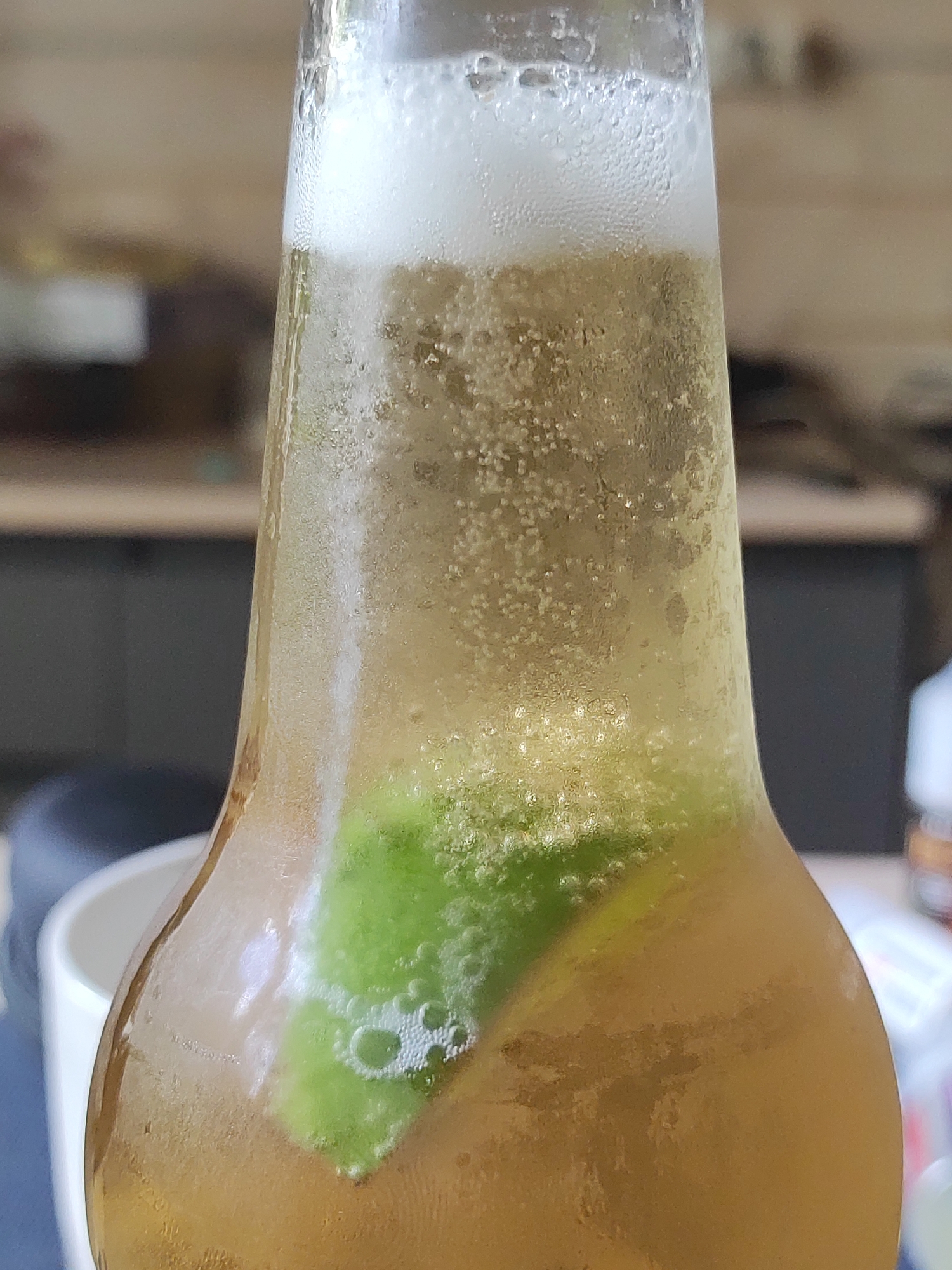 A close-up, slightly out-of-focus shot shows the top of a glass bottle containing a light-colored, bubbly beverage with a slice of green lime submerged within. The liquid is effervescent, with numerous small bubbles rising to the surface, where a frothy head of white foam has formed. The bottle itself is condensation-covered and made of clear glass, showing the amber hue of the liquid. The background is blurred, suggesting an indoor setting with what appear to be kitchen cabinets or shelves. There is no discernible location information within the image that would indicate the city of Alabino, Russia.