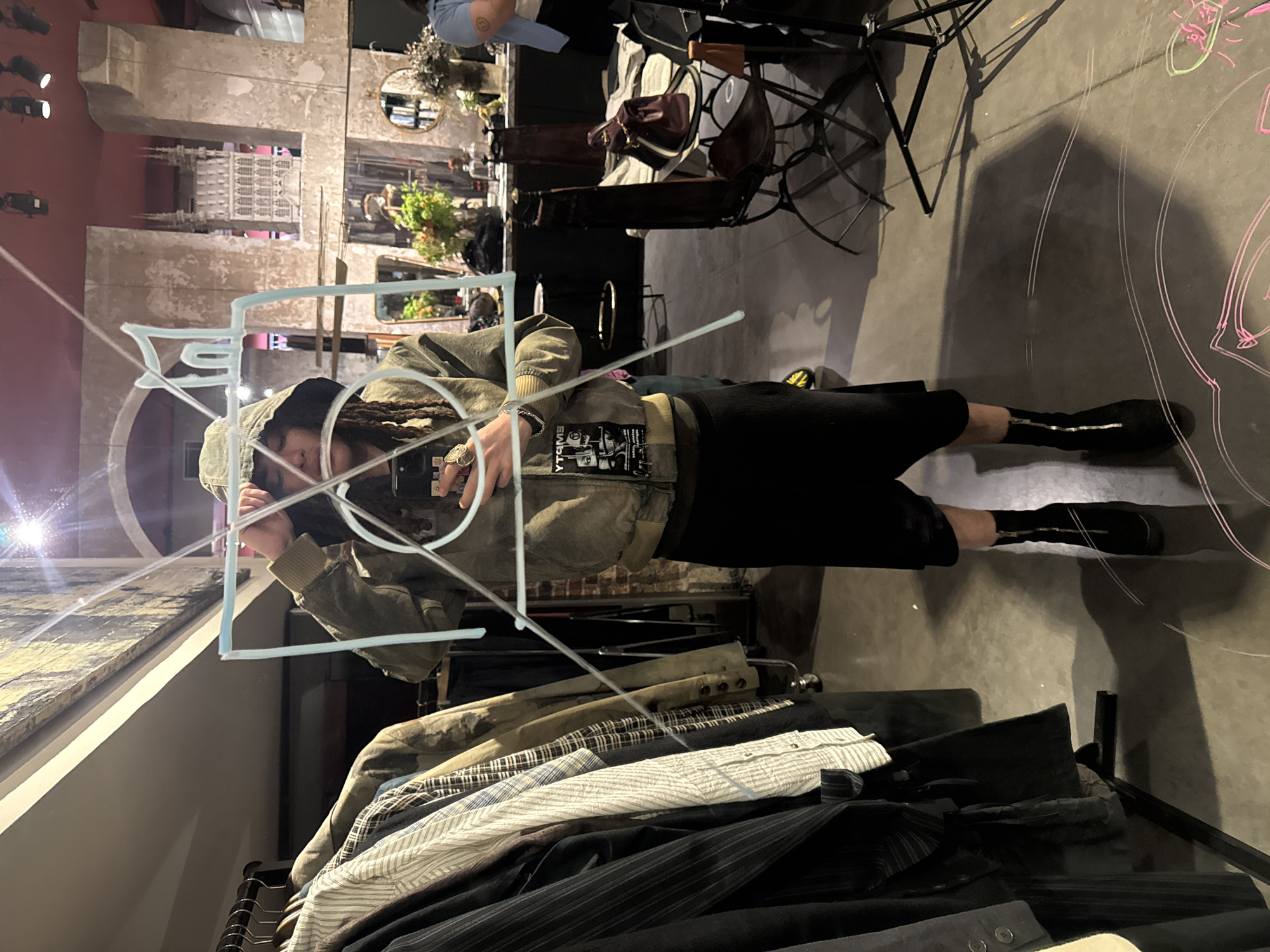 The photograph depicts a person with dreadlocks, wearing a jacket and shorts, standing in front of a reflective surface. The person is holding a phone and appears to be taking a selfie, with a geometric shape seemingly superimposed on the reflective surface. In the background, there are racks of clothing and furniture, suggesting the setting might be a store or showroom. The reflection in the surface shows a cityscape with an archway and detailed architecture, reminiscent of Paris, France. Chalk drawings can be observed on the reflective surface.