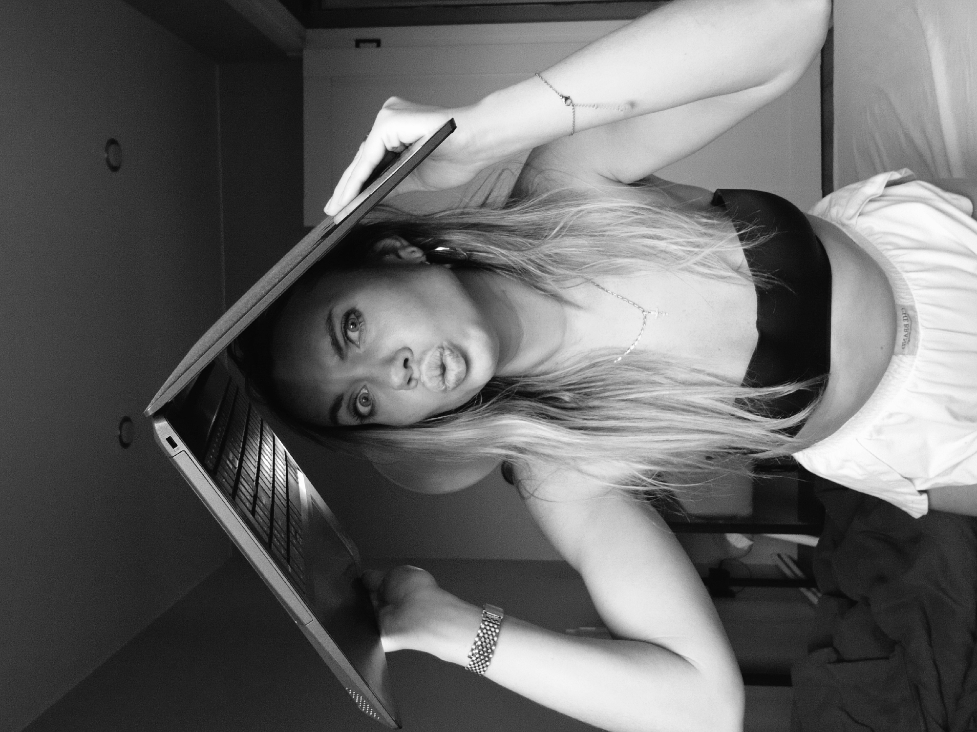 A grayscale medium shot depicts a woman holding a laptop. She is lying on her back, facing the camera with her head tilted towards the left. Her arms are raised, supporting the laptop above her face. She has long, light-colored hair. She is wearing a black top and white bottoms. Her expression includes pursed lips. A bracelet adorns one wrist and a watch the other. A necklace is visible around her neck.
Behind her, a bed and wall are partially visible. The location of the image is not discernible as being in Votkinsk, Russia. No identifying elements are visible.