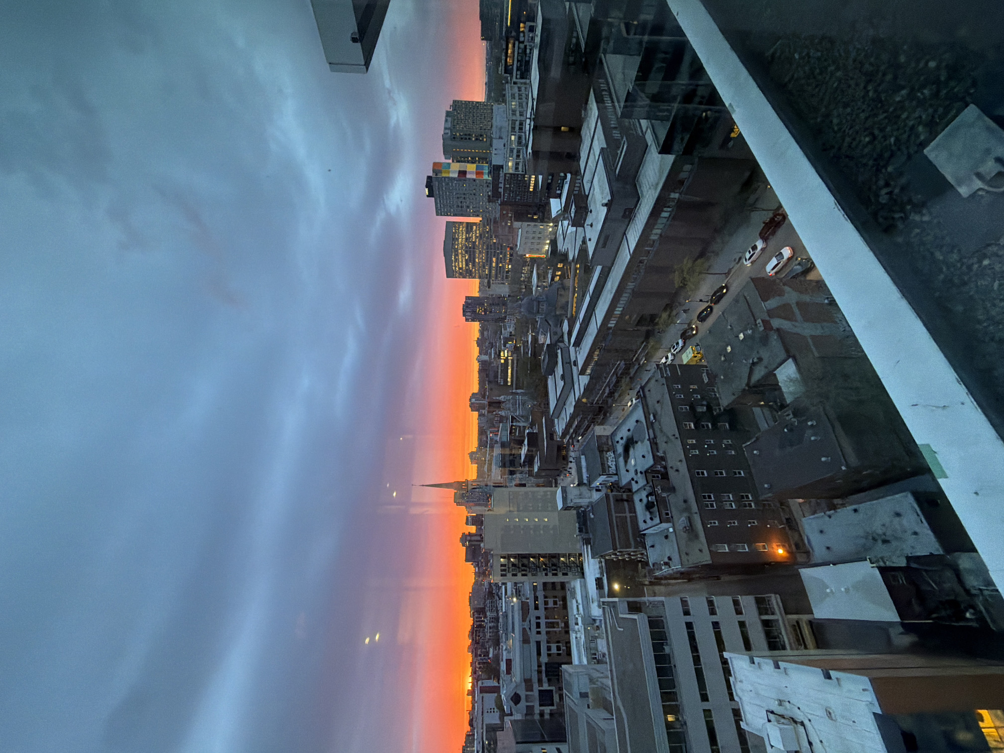 The image captures a dusk cityscape from an elevated vantage point, likely within a building. The sky exhibits a gradient of colors, transitioning from a light, cloud-covered gray at the top to a vibrant orange and red horizon. The setting sun casts a warm glow on the buildings.
Below the horizon, a dense urban landscape stretches out, consisting of buildings of varying heights and architectural styles. A church spire is visible among the buildings. Street lights and illuminated windows are visible. A road runs alongside a row of buildings, on which cars are parked. The image is taken from inside a building and includes part of the wall and window pane of the building.
Based on the buildings and architecture, it appears to be Montreal, Canada. The tall building with the colourful panels is located in Montreal. The church spire visible in the city skyline is likely St. Patrick's Basilica which is located in Montreal.