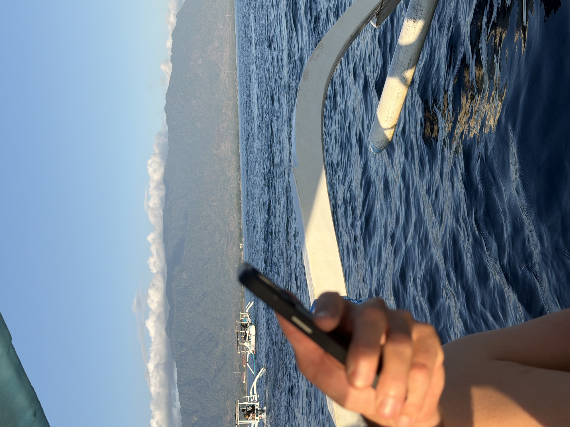 A hand holds a black smartphone in the foreground, obscuring a portion of the frame and its owner's leg. Beyond the hand, the choppy, dark blue water of the sea stretches towards a distant shoreline. In the middle ground, two traditional outrigger boats with white hulls and bamboo outriggers navigate the water. The larger boat in the background appears to have passengers. The background is dominated by a hazy, green mountain range under a clear blue sky with scattered white clouds. The image appears to be taken from another boat, with a portion of its white hull visible in the lower right and a turquoise awning in the upper left. The scene suggests a daytime excursion on the water, possibly in Indonesia, given the boat style.