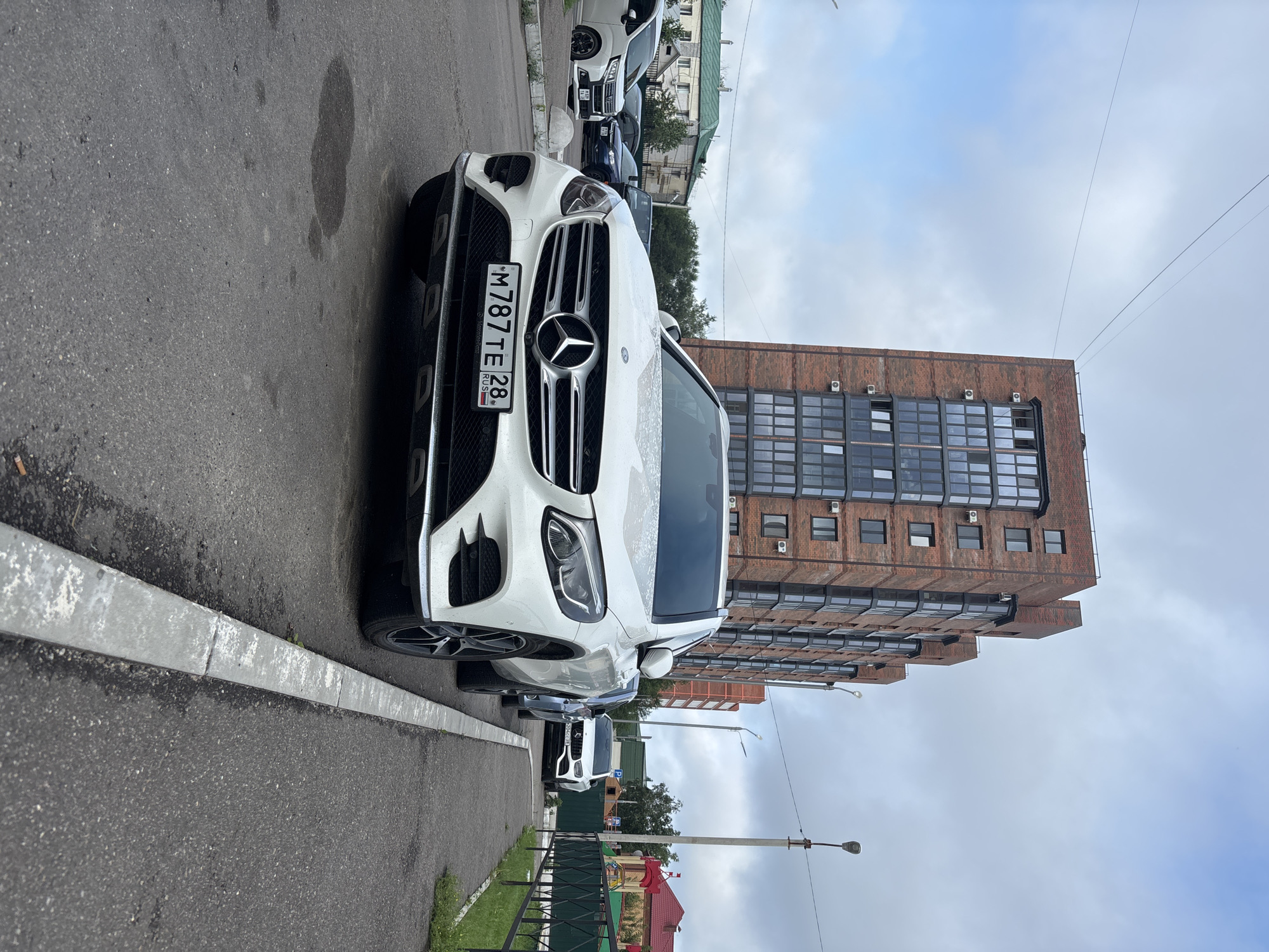 The image depicts a white Mercedes-Benz GLC parked on an asphalt street in Blagoveshchensk, Russia. The car is positioned in the foreground, facing the camera, with its license plate clearly visible: M787TE28 RUS. Behind the car, a multi-story brick apartment building dominates the background, featuring numerous windows and balconies. To the right of the car, a sidewalk runs alongside a green fence that encloses a playground area with a red structure visible. Other vehicles are parked further down the street, including a white compact car and a dark-colored SUV. The sky is overcast with scattered clouds. The scene suggests a residential urban environment on a cloudy day.