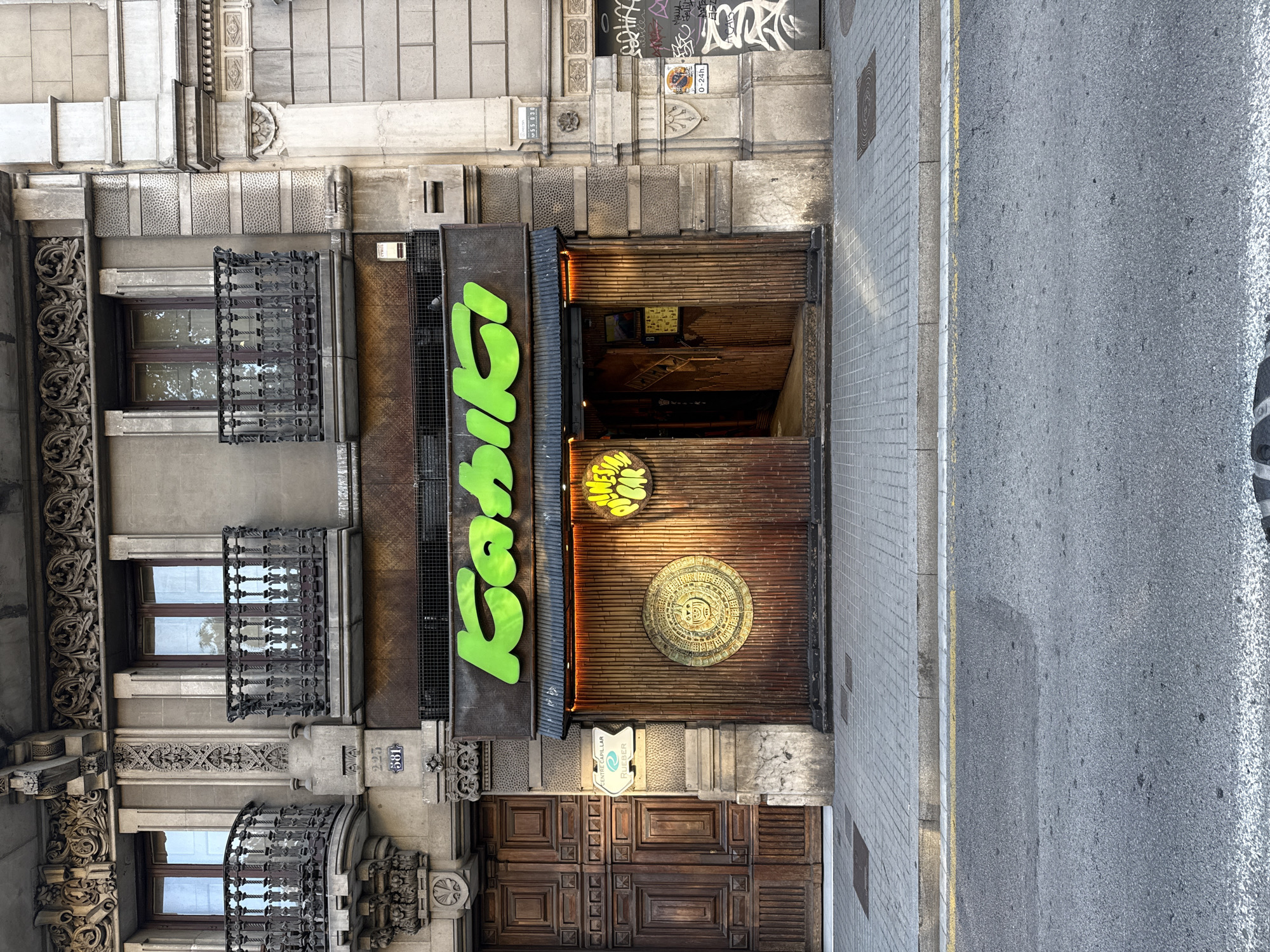 This is an outdoor, eye-level, medium shot of the facade of a building with the restaurant "Kaktus" (cactus in Catalan), located in Barcelona, Spain.
The building has a stone facade with ornate details around the windows, which have small wrought iron balconies. The restaurant's signage is vertical and in bright green, the letters "Kaktus" are stacked on top of each other.
The restaurant's entrance is a dark wood and bamboo design, with a large Aztec-inspired circular design in the center. Above the entrance is a smaller sign for "Mines Public". Below the Aztec design is a sign that says "Centre Capilar Rieber".
To the right of the building is a sidewalk and a road. There are no people visible in the image.