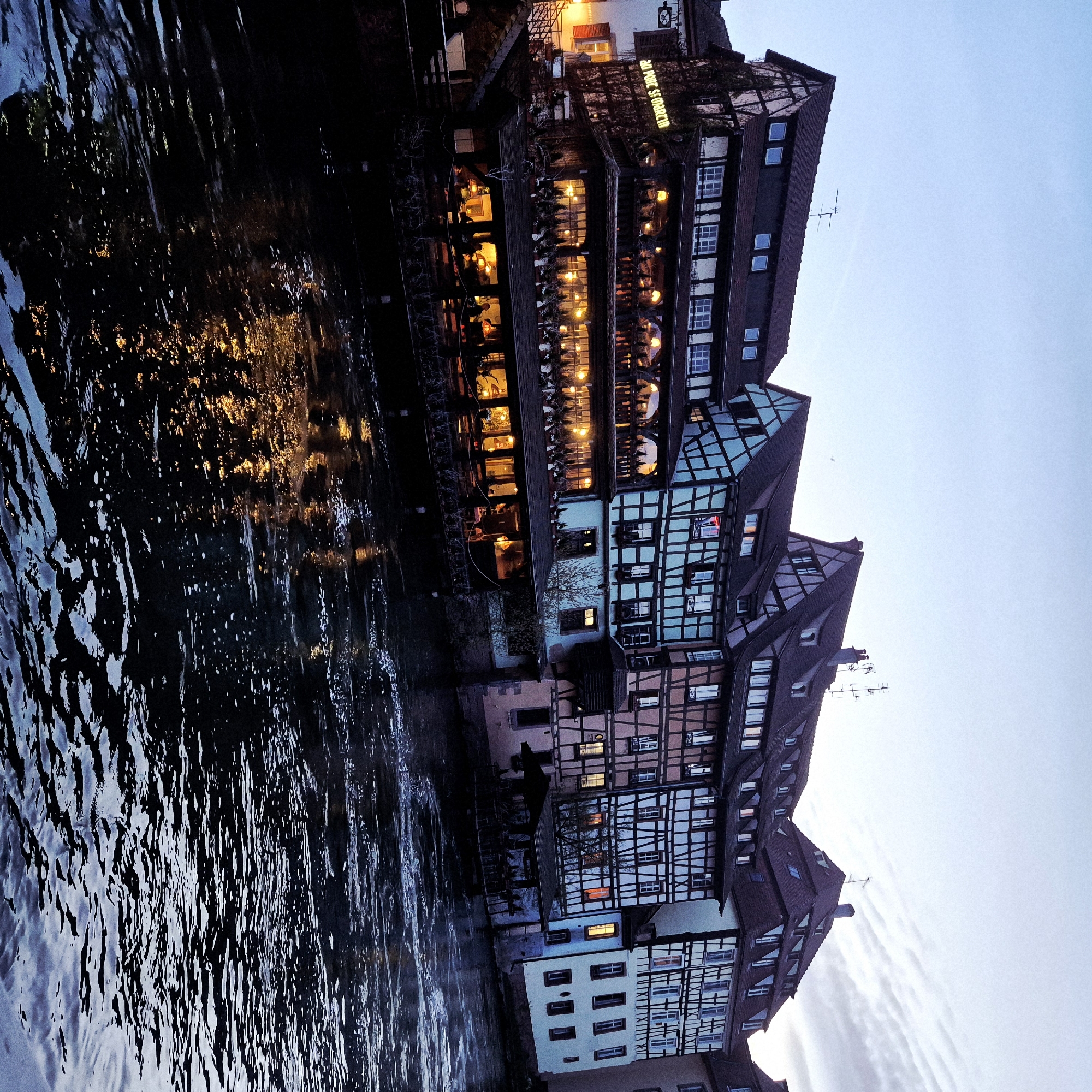 This image depicts a twilight scene in the Petite France district of Strasbourg, France, along the Ill River. A series of historic half-timbered buildings with steeply pitched dark roofs line the riverbank.
The prominent structure on the left is a multi-story building, identified by a glowing sign as "AU PONT ST MARTIN." This building functions as a restaurant or establishment with multiple illuminated dining levels extending over the dark, rippling river. The lowest level features an enclosed or covered patio area directly above the water, where numerous indistinct figures are visible, seated at tables under warm lighting. Above this, a second story also shows patrons through large windows, with additional potted plants lining its exterior ledge. The building's facade is partially covered in dark ivy, contrasting with its lighter half-timbered sections.
To the right of "AU PONT ST MARTIN," a sequence of other half-timbered houses continues along the river. These structures display varied pastel facade colors, including light blue, pink, and white, all framed by dark timbering. Windows in these buildings are intermittently lit, casting warm light. Rooftop antennas and chimneys are visible against the soft blue-grey sky, which features subtle cloud streaks. The river in the foreground is dark, reflecting the artificial lights from the buildings as elongated, shimmering streaks of yellow and orange, interspersed with areas of white foam. The water appears to be flowing, creating ripples and texture across its surface.