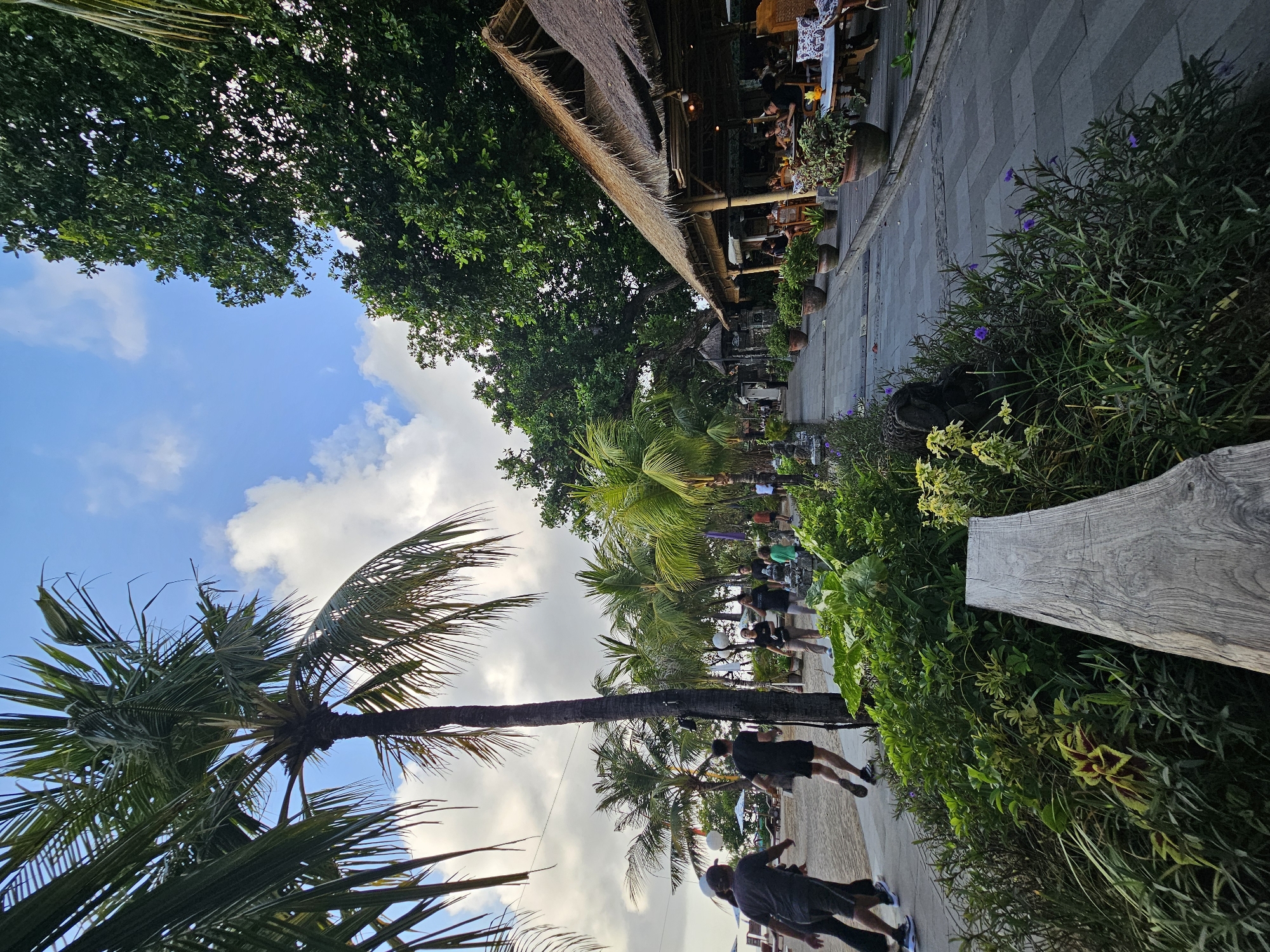 The image depicts a beachside promenade in Denpasar, Indonesia, likely near Seminyak or Kuta. The scene captures a pathway with paved tiles, bordered by lush green vegetation and potted plants. Several people are walking or running along the path. To the right, a beachfront restaurant with a thatched roof is visible, with patrons seated at tables. Palm trees line the left side of the pathway, with the sandy beach and ocean visible in the background, under a cloudy sky. The overall ambiance suggests a casual, tropical setting with a mix of natural beauty and developed tourist infrastructure.