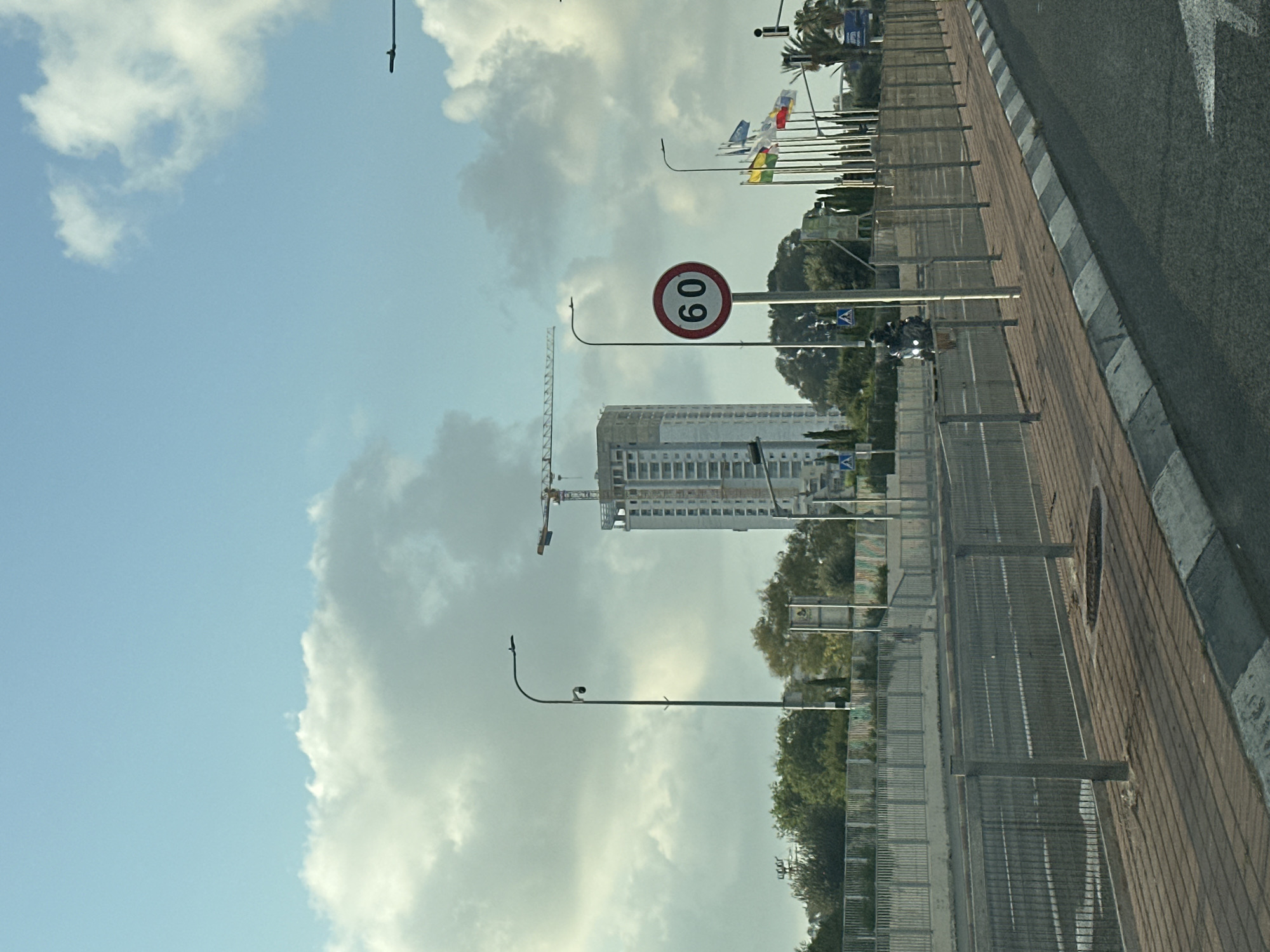 This image captures a street scene in the city of Mate Asher Regional Council, Israel. A multi-story building under construction, complete with a crane, dominates the background. In the foreground, a road with a sidewalk runs alongside a metal barrier. A speed limit sign displaying "60" is visible on a pole. Several flags are also planted along the roadside, and a motorcycle is seen moving on the road. The sky is partly cloudy.