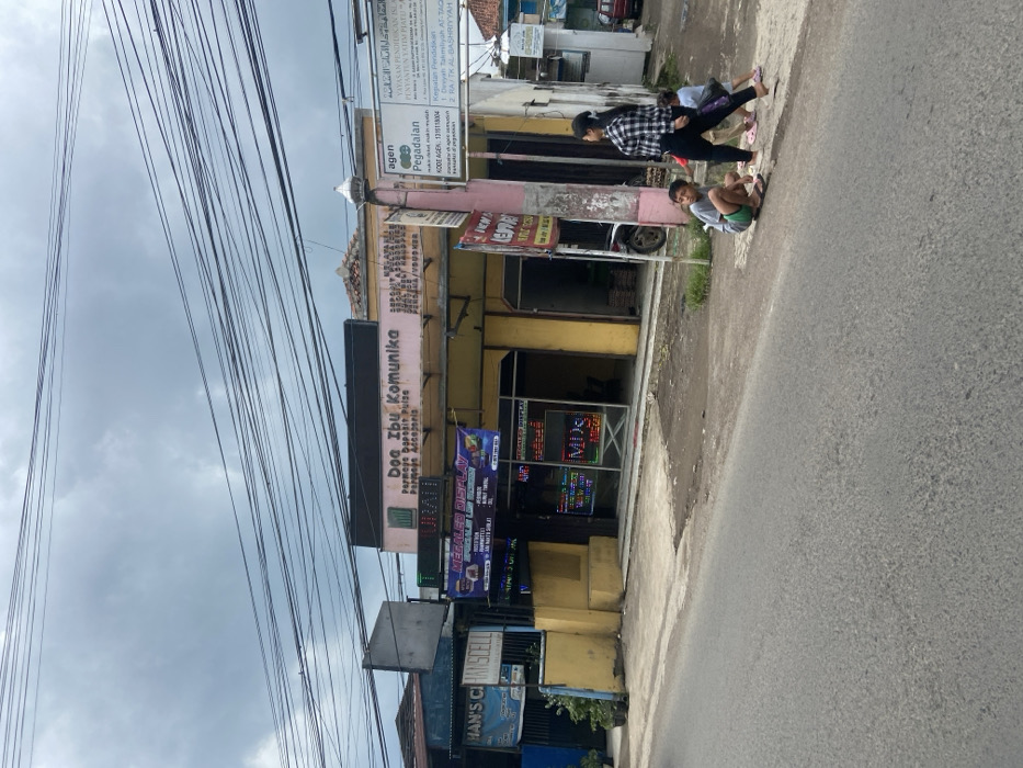 The image shows a street corner in Ciparay, Indonesia (latitude: -7.023, longitude: 107.687). The scene is a typical residential area with a small commercial storefront on the corner. The storefront is called "Dog Ibu Komunik" and is likely a cell phone shop. The building has a yellow facade with a large sign above the door. To the right of the shop is a pole with signs and advertisements for "agen pegadalan" and a foundation called "Yayasan Pendidikan". Two children, one boy and one girl, are walking in the street, with the boy appearing to be looking at something on the ground. An adult woman with a grey blouse and a black skirt is walking behind them. The street is paved with asphalt and appears to be relatively narrow. There are many power lines and telephone wires overhead, indicating the presence of urban infrastructure. The sky is overcast with clouds, suggesting the possibility of rain. There are a few cars and motorcycles parked on the side of the street. The scene is somewhat deserted, but it is clear that this is a neighborhood where people live and work.