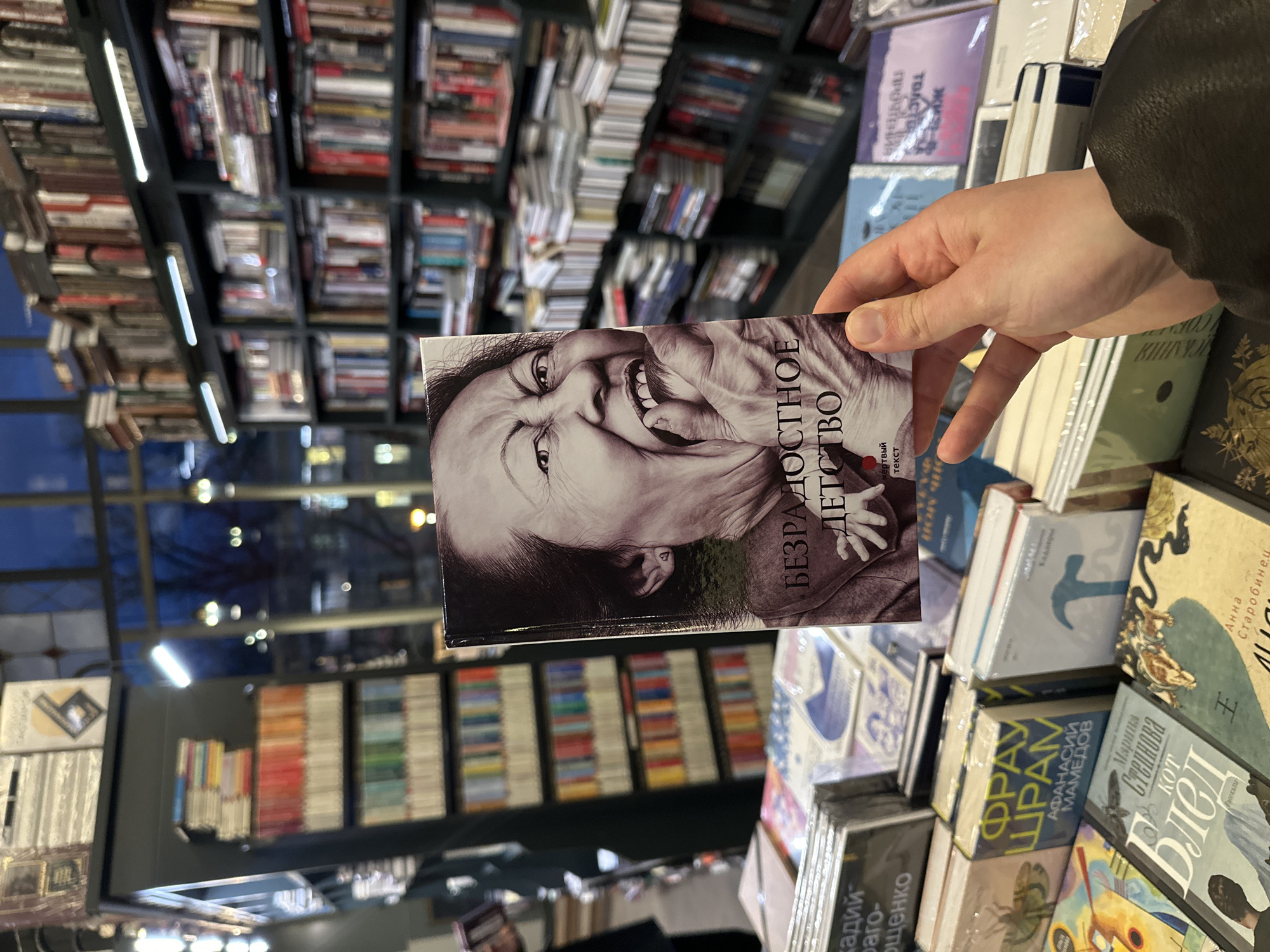 The image shows a hand holding a book titled "Безрадостное детство" (translated: "Joyless Childhood"), with a black-and-white portrait of an Asian woman on the cover. The book is held in a bookstore, with shelves filled with other books visible in the background. The location appears to be a bookstore in Saint Petersburg, Russia. The hand holding the book belongs to a person wearing a dark jacket. The foreground contains stacks of other books for sale, some still wrapped in plastic. Other books stacked at the bottom say “Фрау Шрам” (translated: “Frau Scar”), “Марина Степнова Кот Блед” (translated: “Marina Stepnova Cat Pale”) and "Анна Старобинец ВИСКАНИЯ" (translated: "Anna Starobinets VISKANIYA”).
