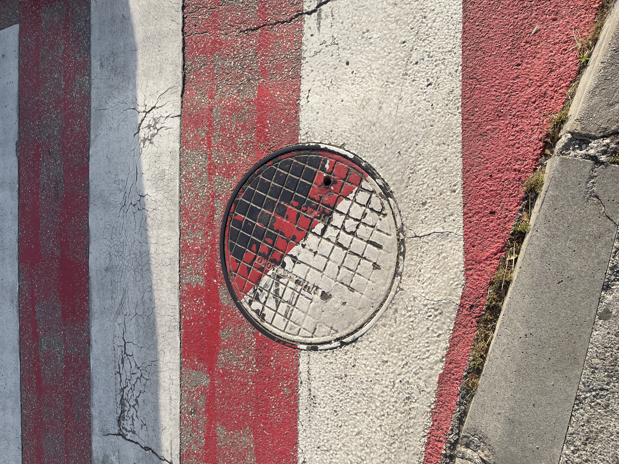 A top-down view captures a section of a crosswalk in Budva, Montenegro, featuring alternating red and white stripes painted on asphalt. A circular manhole cover, partially painted with red and black squares on one side and white on the other, disrupts the stripes in the center of the frame. Cracks and wear are visible in the painted asphalt, indicating age and use. The edge of a concrete curb runs along the right side of the frame, with grass and debris accumulating between the curb and the painted crosswalk. No people are visible in the shot. The overall lighting is bright and even.