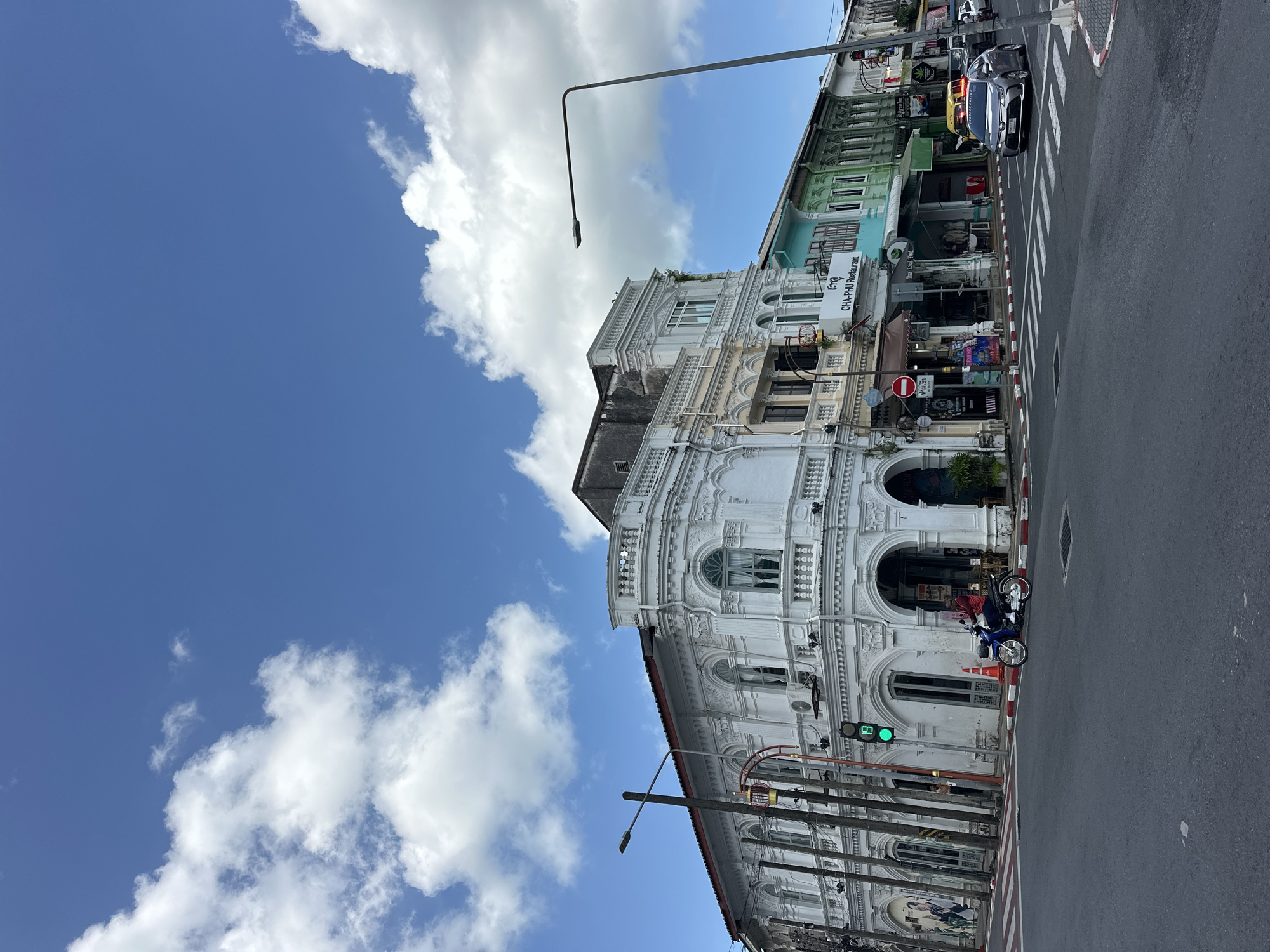 The image depicts a street scene in Phuket City Municipality, Thailand, characterized by Sino-Portuguese architecture. A prominent white building with intricate decorative elements and arched doorways stands on the left, forming a corner. Adjacent to it, a row of colorful buildings, painted in pale yellow, turquoise, and green, continues along the street. The sky above is a vibrant blue with scattered white clouds.
In the foreground, a street intersection is visible with pedestrian crosswalks. A motorcycle with a rider wearing a red shirt is stopped at a traffic light, which is currently displaying a green signal. Several utility poles and a street lamp are present. Cars are visible on the road, including a silver sedan crossing the intersection. Signs are posted, including one for "CHA-PHU Restaurant." The overall atmosphere suggests a sunny day in an urban environment.