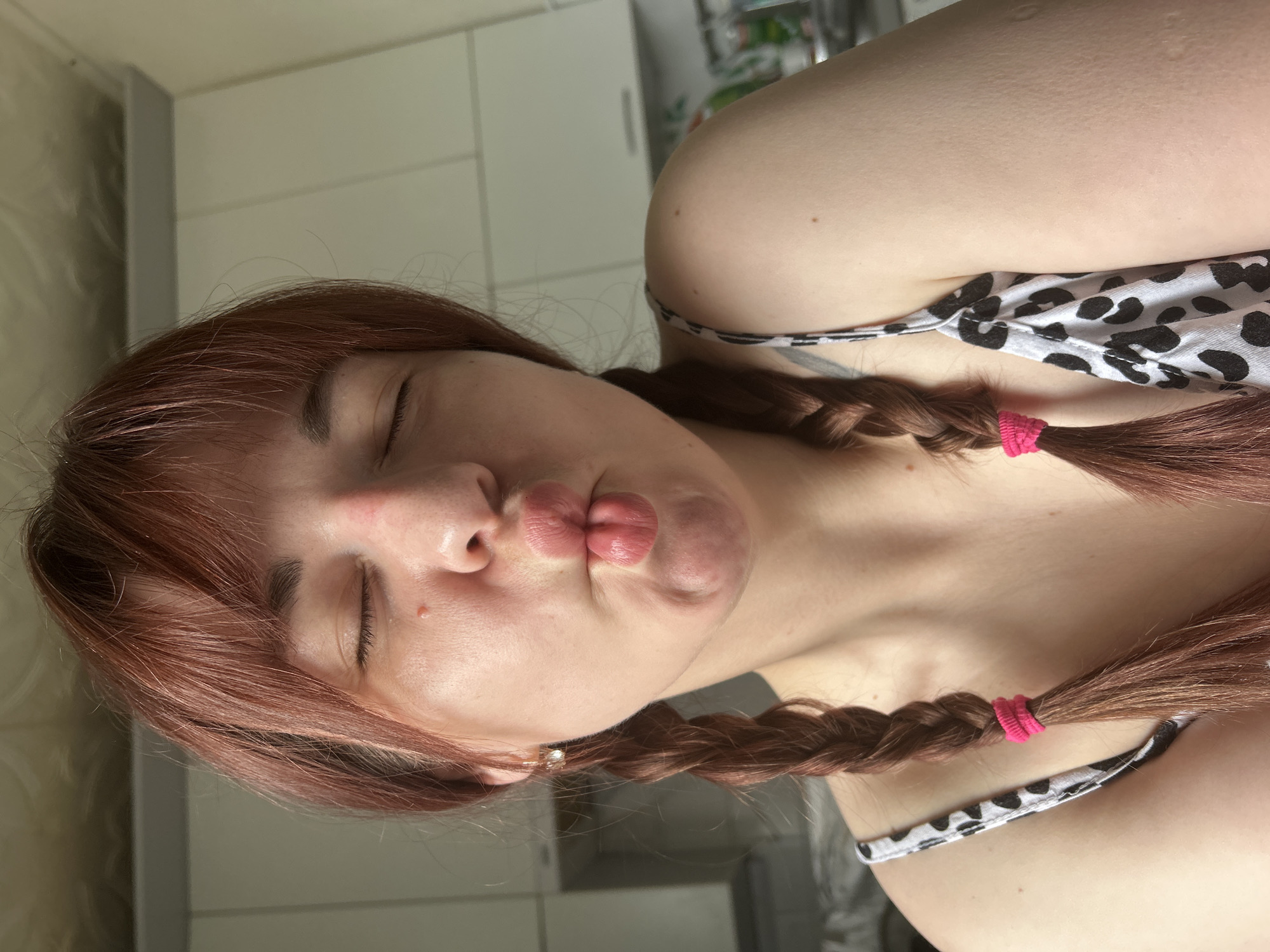 A woman with auburn hair styled in two braids with pink elastics is pouting her lips toward the camera. Her eyes are closed. She is wearing a white tank top with a black Dalmatian print. The background shows a white wall, possibly in a bathroom or kitchen, and a blurred white cabinet. The image shows no indication of being in the city of Фоки, Russia.