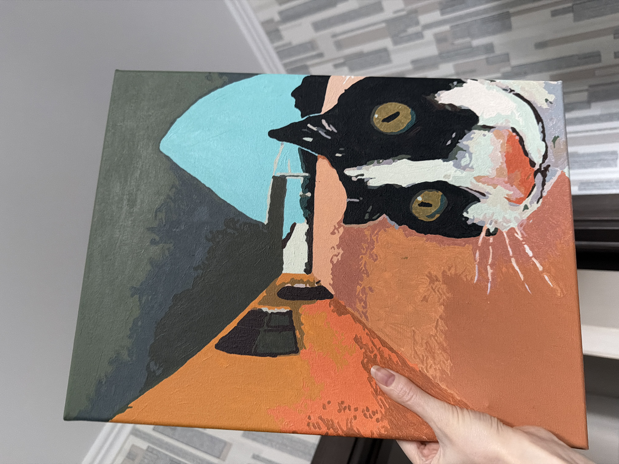 A person's light-skinned hand holds up a rectangular canvas painting, likely depicting a stylized cat. The painting features a large, abstract rendering of a cat's head, appearing upside down or tilted. The cat is primarily black and white, with striking golden-yellow eyes that have black pupils. There are also patches of light orange, light blue, and grey within its fur. Several thin white lines suggest whiskers or fur details emanating from the cat's face.
The background of the painting is composed of distinct, blocked areas of color. On the left, there's a dark greenish-grey section that transitions into a bright, almost sky-blue shape. The majority of the lower and right parts of the background are filled with various shades of orange and brown, creating a sense of perspective and depth, possibly resembling a corridor or a room with architectural elements like doorways or windows in dark brown/black shapes. The painting's texture suggests it might be acrylic on canvas, possibly from a paint-by-numbers kit due to the clear segmentation of colors.
The hand, with short, unpainted fingernails, cradles the bottom and right edges of the canvas. The background of the photograph shows indoor setting with a patterned wall featuring grey and white rectangular tiles in the upper right, and a plain grey wall on the left. The scene is brightly lit. There is no visible text in the image.