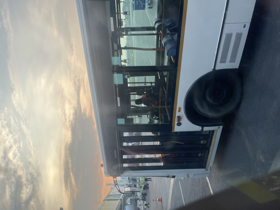 The image, correctly oriented, depicts the side view of a white airport apron bus parked on the tarmac of an airport, likely in Guwahati, India. The sky above is illuminated with soft, warm hues of orange, yellow, and grey, characteristic of either dawn or dusk, indicating the "golden hour."
The bus features large windows revealing its interior, where blue seating and a driver figure are discernible. Reflections in the glass suggest other passengers or objects inside. A prominent black rear tire of the bus is visible. The bus's front door is open, suggesting boarding or disembarking activity.
Adjacent to the bus's front, parts of a jet bridge structure and various pieces of airport ground equipment, such as ladders and an orange cone, are visible. Several ground crew members, some in high-visibility vests, are seen around the bus and the jet bridge, indicating active airport operations. The tarmac is dark asphalt.
Visible text includes "EMERGENCY EXIT" written in white on a red background inside the bus window. Other smaller texts on the bus's exterior panels are present but unreadable. The overall scene captures a moment of passenger transfer or preparation at an airport apron under clear and calm weather conditions.