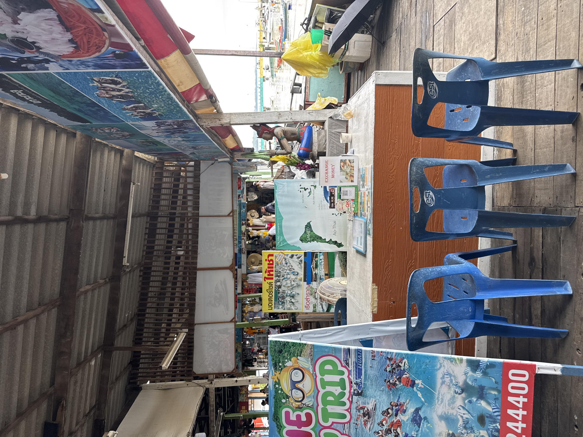 The image depicts a waterfront stall in Ban Phe, Thailand, likely a tourist information booth or souvenir shop. The stall is constructed of weathered wood and covered by a corrugated metal roof. Directly in front of the stall are three identical blue plastic chairs. Behind the chairs, a wooden counter displays various promotional materials, including posters advertising tourist activities like boat trips and jet skiing, as well as a map of local islands. One poster features the Thai text "มอเตอร์ไซค์ ให้เช่า" which translates to "Motorcycle for rent". Another poster is an "EXCHANGE MONEY" sign. A small Buddha statue sits on the counter, adorned with a yellow sash and flowers. A bright yellow plastic bag is prominent on the right side of the scene, along with a bicycle and various other objects suggesting a working commercial space. In the background, boats are moored in the water, with a distant bridge visible. The overall impression is one of a bustling, casual tourist hub.