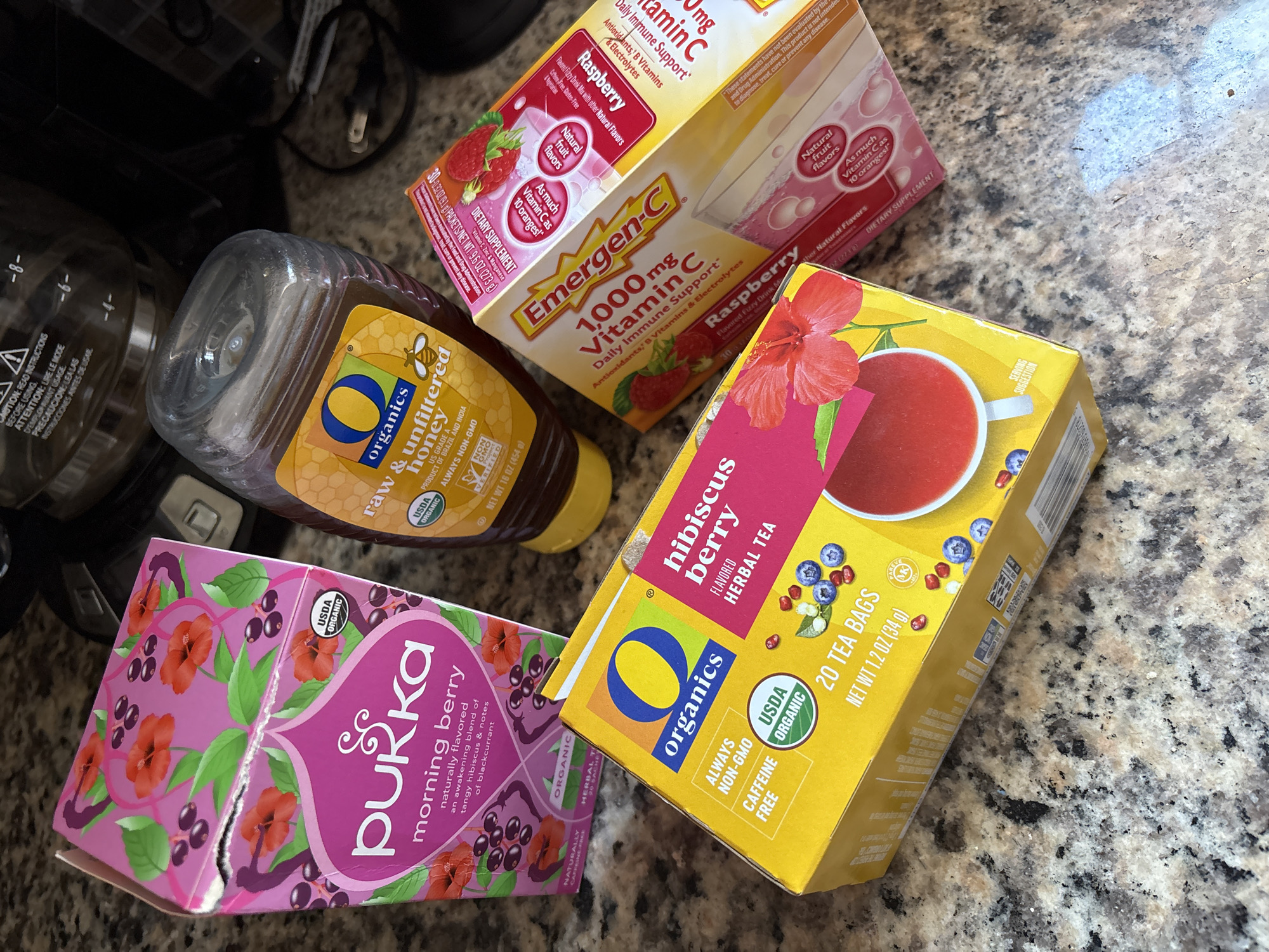 A high-angle shot captures a collection of health and wellness products on a countertop with a mottled beige and brown pattern, possibly granite. In the top left corner, the edge of a black coffee maker is visible with its power cord snaking down.
The items present, clockwise from the top, are: a box of Raspberry Emergen-C 1000mg Vitamin C packets; a box of "O Organics" Hibiscus Berry Herbal Tea containing 20 tea bags; an "O Organics" raw and unfiltered honey bottle; and a torn, partially crushed box of Pukka Morning Berry tea.
The setting appears to be a residential kitchen. There are no visible people or direct interactions.