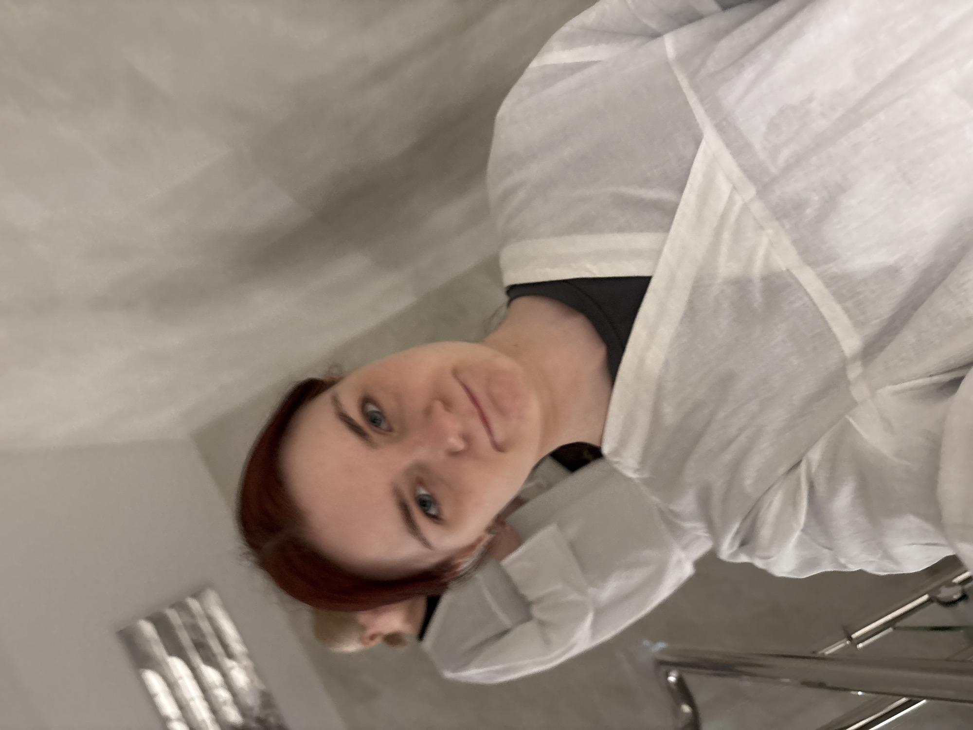 In a dimly lit, indoor setting, two individuals are present, both wearing white disposable smocks over dark clothing. The foremost individual, a woman with red hair, faces the camera, her expression neutral as she looks directly into the lens. Her eyes are open, and her mouth is closed. Behind her, another person, appearing to be a woman with blonde hair, is visible from the back, also wearing a white smock. The background is largely out of focus, with a tiled wall and a metallic railing visible. There is no indication of the specific location in Saint Petersburg, Russia.