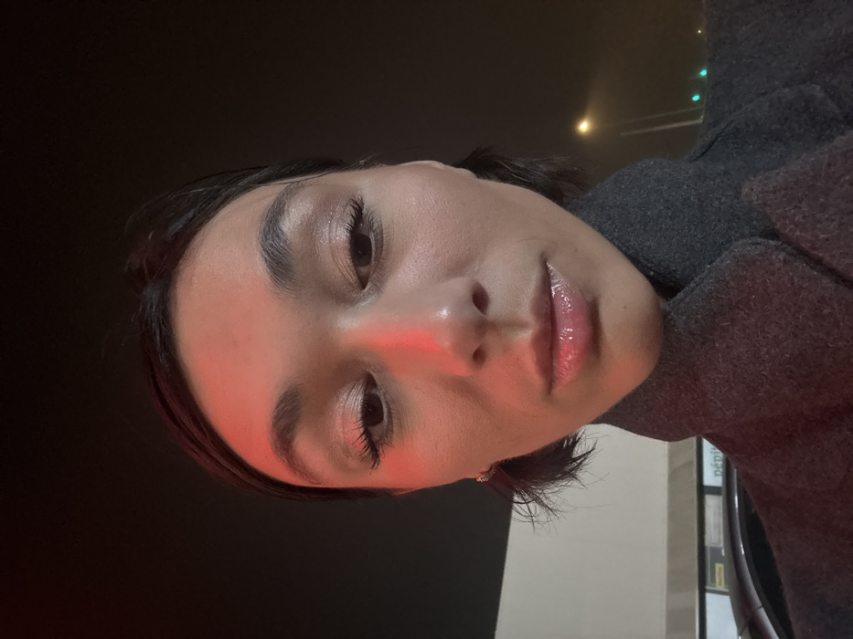 A person with light skin, dark hair, and glossy lips faces the camera at an angle. The lighting is dim, with a reddish hue on the nose and forehead, likely from an external light source. The individual is wearing a gray wool coat or jacket. The background appears to be a dark, possibly nighttime, setting. Hints of street lights and building elements are visible in the background. The person's expression is neutral.
