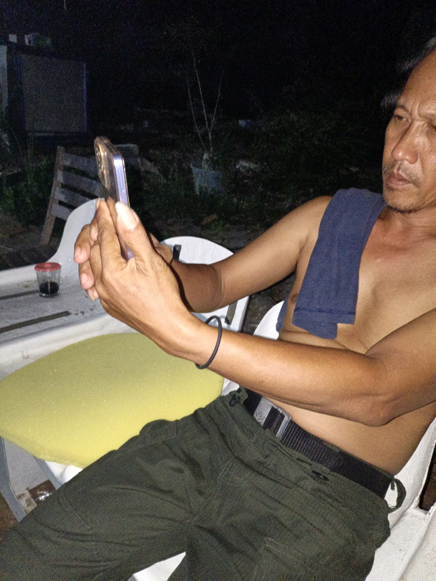 The image shows a middle-aged man sitting outdoors at night. He is wearing dark green cargo pants and no shirt, with a dark blue towel draped over his shoulder. He is holding a smartphone in his right hand and appears to be looking at it. In front of him is a white table with a yellow cushion on one side and a small red-lidded container of dark liquid on the other. The background is dark and out of focus, suggesting a rural or semi-rural setting at night, possibly in Cikal, Indonesia. There are some plants and structures visible in the background.