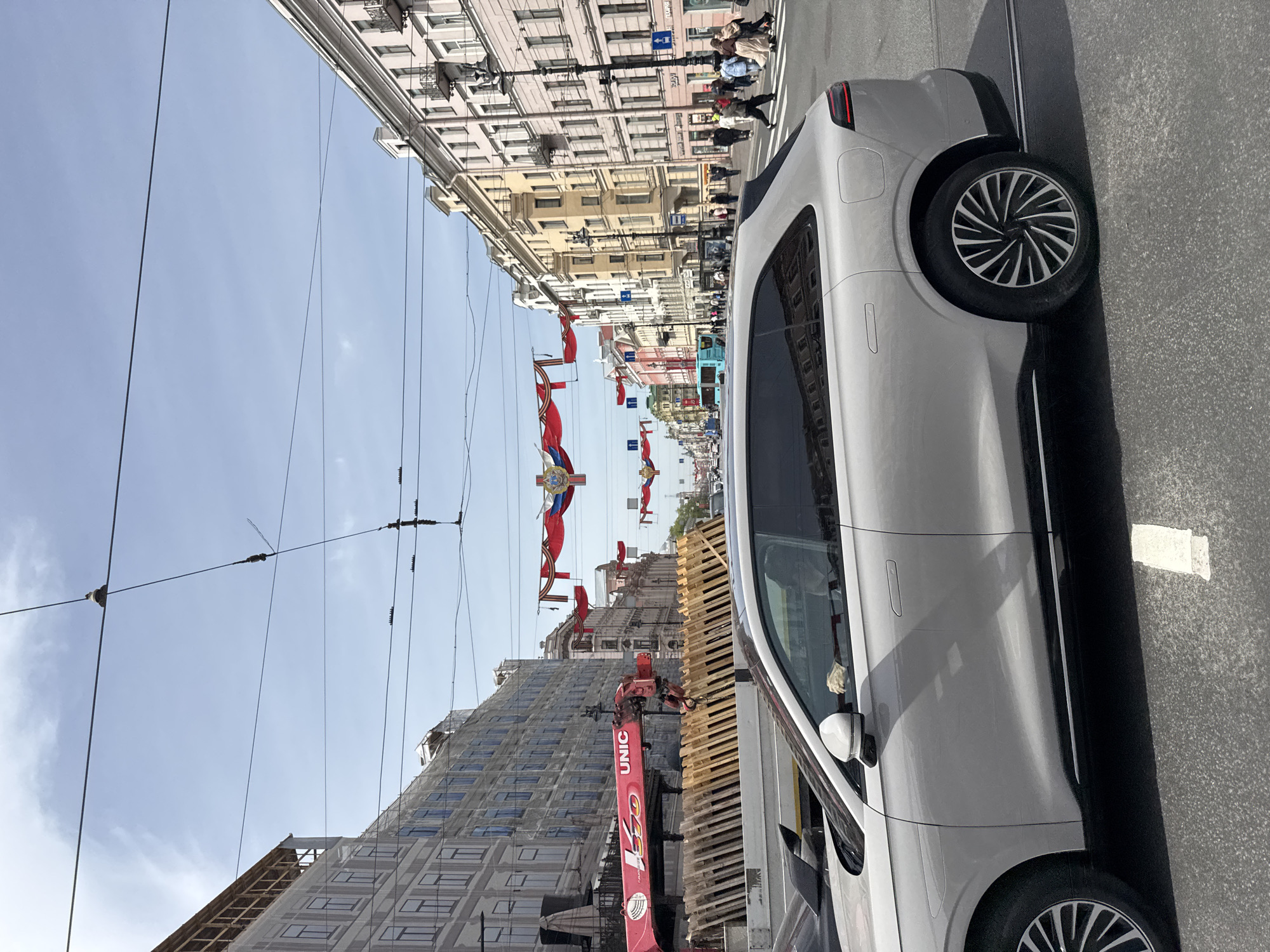 A street-level, slightly angled shot shows a street in Saint Petersburg, Russia. A light-colored vehicle occupies the right side of the frame. The street stretches into the distance, lined with buildings. Red banners hang across the street, displaying a star and wreath symbol. People are visible in the distance, some walking on the sidewalks and others crossing the street. A building under construction with scaffolding and a crane is visible on the left side of the street. Overhead wires traverse the sky. The weather appears clear and sunny.