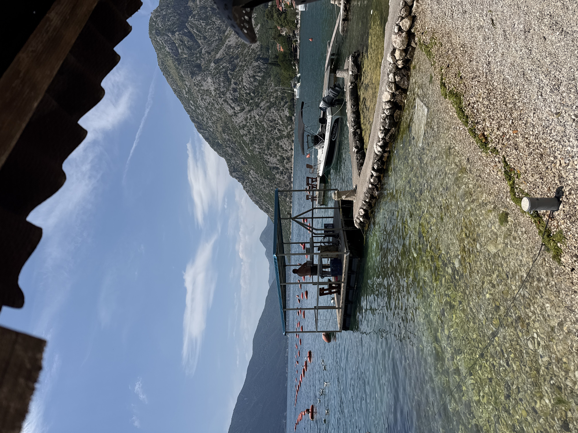 An outdoor daytime scene at a waterfront location in Donji Orahovac, Montenegro, captures a picturesque view of a bay or inlet. The water is clear and shallow near the shore, revealing a rocky seabed, transitioning to a deeper blue further out. A man stands on a floating platform, possibly engaged in aquaculture or maintenance, under a blue-covered canopy. The platform is equipped with tables and chairs. Surrounding the platform, numerous orange buoys dot the water, arranged in rows, suggesting a farming or marking system. Seagulls are present in the water near the platform. In the background, a boat is docked at a pier, and beyond that, a majestic, rugged mountain rises, its slopes covered in sparse vegetation. The sky is blue with scattered white clouds. Part of a wooden roof structure is visible in the upper portion of the frame.
