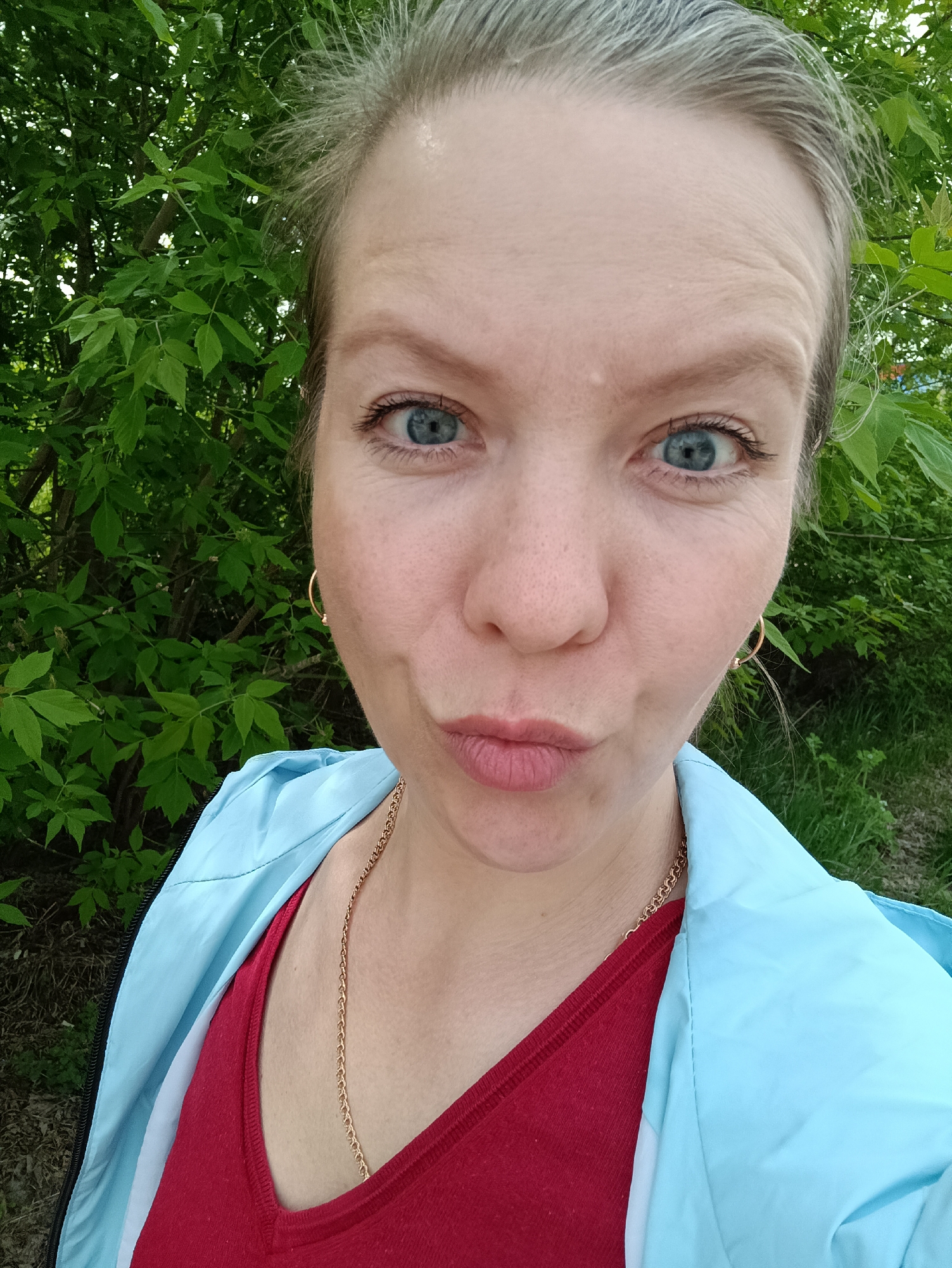 A selfie of a light-skinned woman with light blonde hair, pulled back, wearing gold hoop earrings, a gold chain necklace, a red shirt and light blue jacket. She is in front of green foliage. The location is Chaikovsky, Russia.