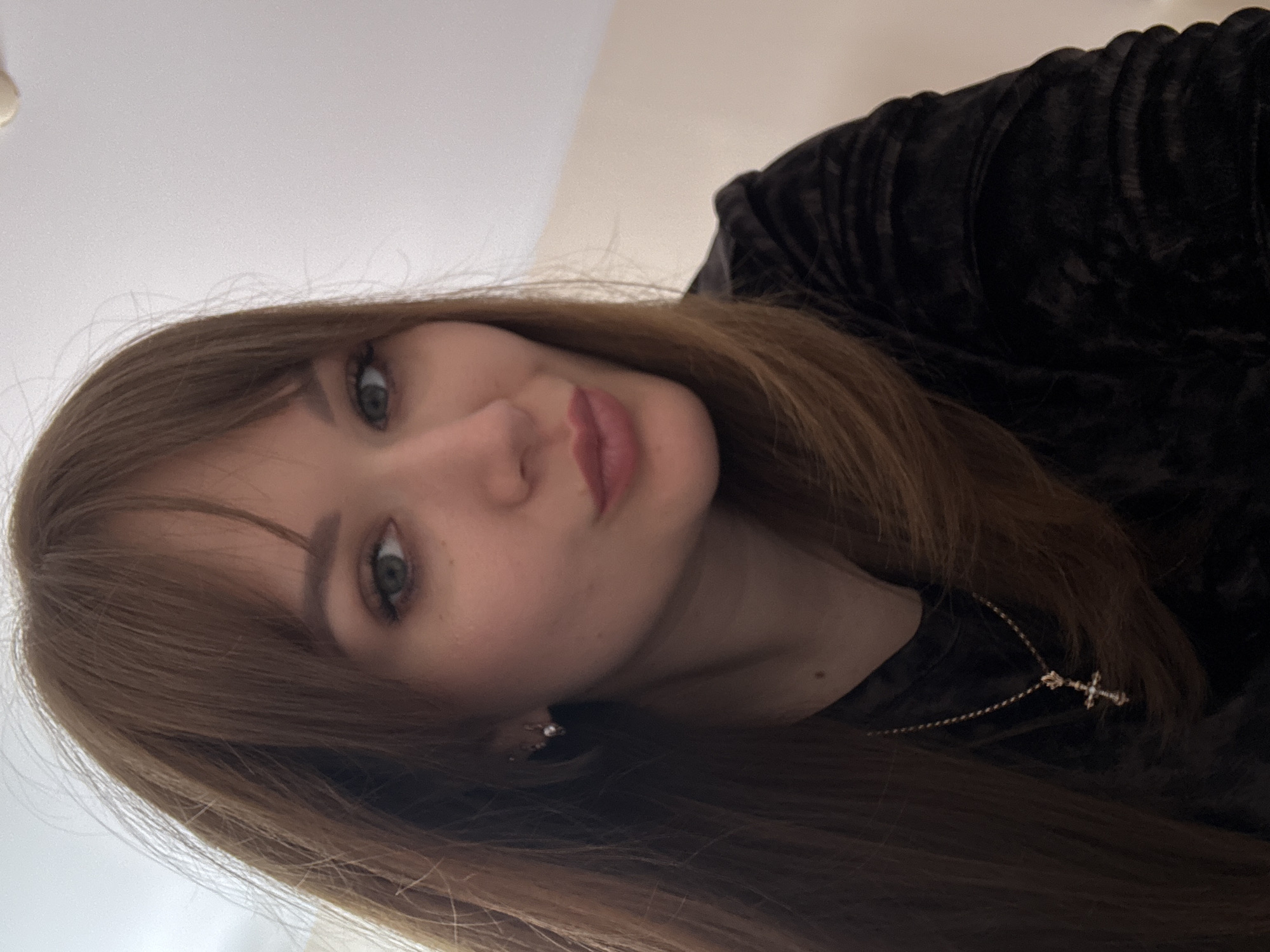 A close-up, angled portrait of a young woman. She has light brown hair with bangs and appears to be wearing a dark, possibly velvet top. A delicate gold necklace with a cross pendant is visible. Her eye color is greenish. The background is out of focus but appears to be a plain light-colored wall or ceiling. The picture does not depict any specific location, people, subjects, objects, actions, interactions, or the environment in the city of Yevpatoriya, Ukraine.