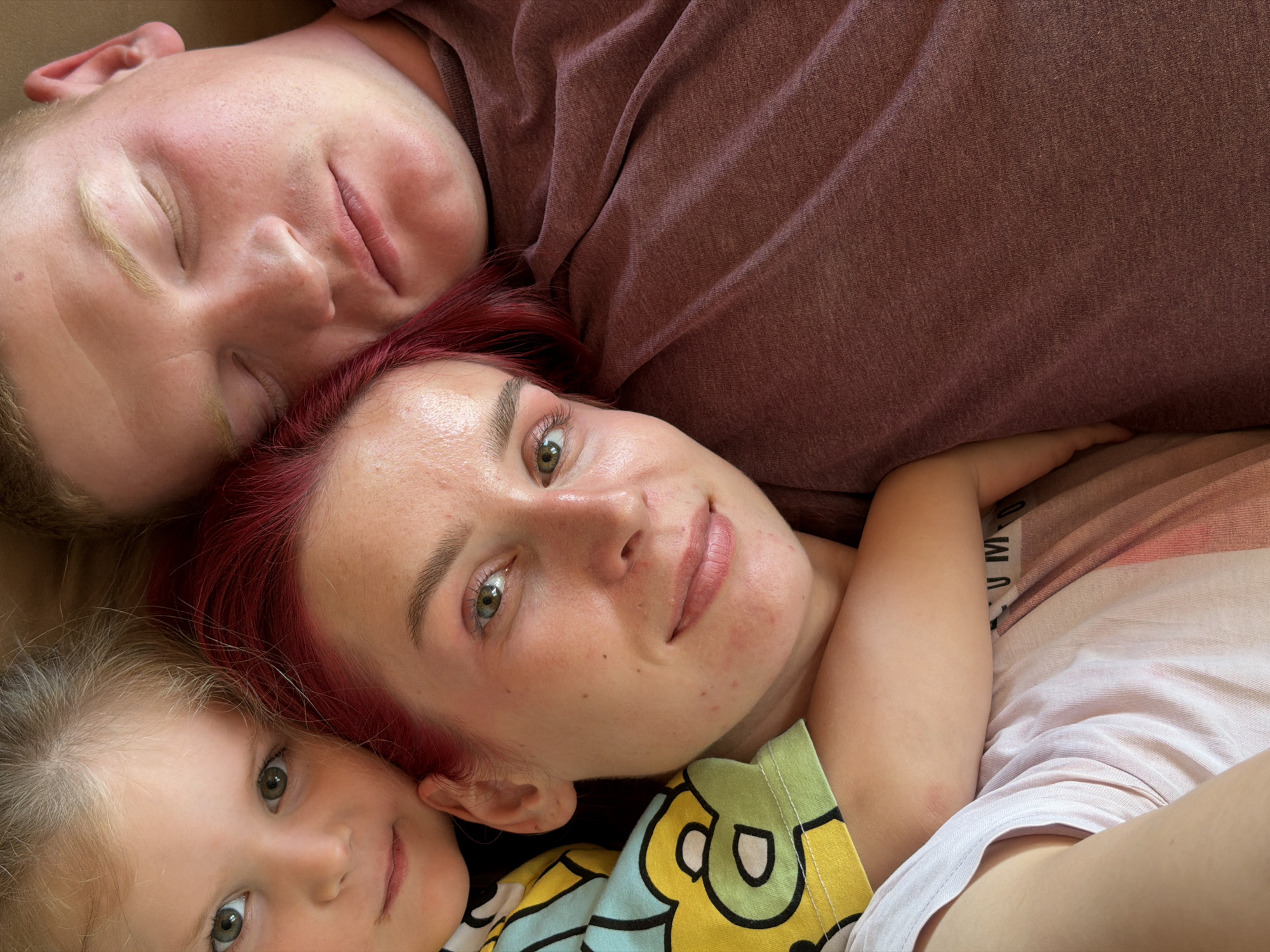 A close-up, top-down shot shows three people lying down, appearing to be sleeping or resting. On the left, a young child with blonde hair and green eyes looks towards the camera. Next to the child is a woman with bright red hair and green eyes, also looking towards the camera with a slight smile. The man is positioned to the right, with his eyes closed and head tilted, resting his head on the woman's shoulder. The background is a textured surface, possibly a bed or couch, with hints of fabric in muted colors like brown, pink, and cream. There is no indication of the city of Chaikovsky, Russia, or any other specific location within the image.