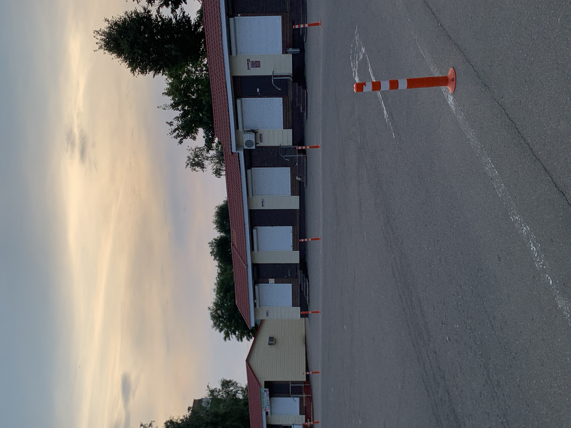 This is a vertical, medium-shot, eye-level photograph of a row of small commercial buildings with red tile roofs, possibly in a semi-rural or suburban area, taken at dusk in Kapyĺ, Belarus. The buildings appear to be single-story structures with light-colored facades and dark brown trim. Each unit has a dark door and a white roller shutter, indicating they are closed. Some of the buildings have air conditioning units installed. In front of the buildings, there is a paved area, likely a parking lot, with several orange and white traffic cones. A white line is painted on the pavement in the foreground, extending from the lower left corner towards the right. Trees are visible behind the buildings, and the sky above is a soft gradient of pale blue and orange, characteristic of sunset. There are no people visible in the image.