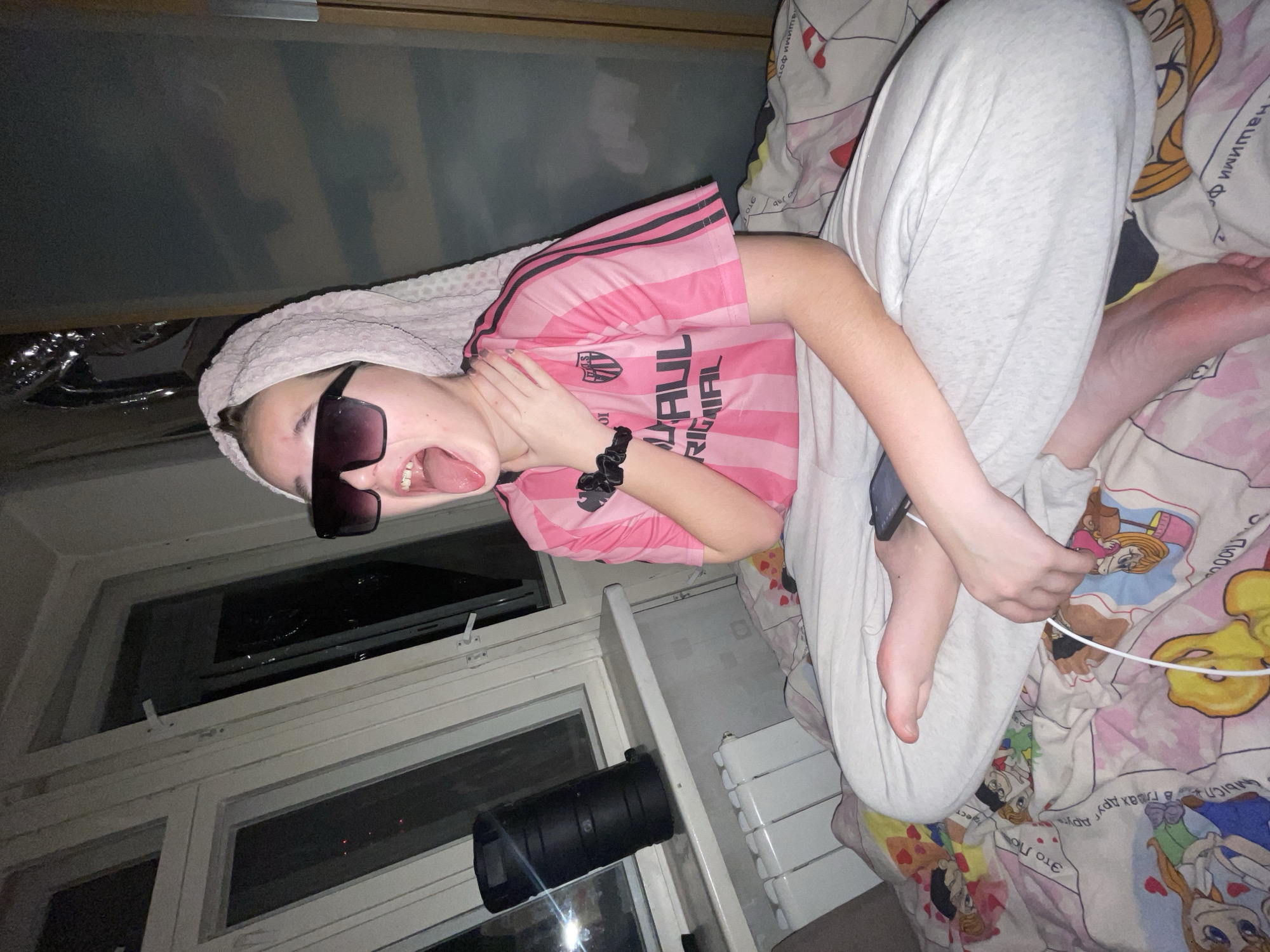 An informal, portrait-style media file, rotated 90 degrees, features a young person lying indoors, likely in a bedroom in Murino, Russia, during nighttime. The subject is seen from the chest up, with their head resting on what appears to be a pillow covered by a white, textured towel. They are wearing dark sunglasses and a pink and black striped athletic shirt, possibly a soccer jersey, with a visible logo and text that includes "RIGAIAL" and "10000" and partially "Нашими фо". A black scrunchie is on their wrist.
The person's expression is playful, with their mouth open and tongue sticking out. Their left hand is raised towards their neck, fingers touching. They are clad in light grey sweatpants, and their bare feet are visible towards the right side of the frame. A smartphone, connected to a charging cable, rests near their feet on the bed.
The bed is covered with a brightly colored duvet or sheet featuring various cartoon characters and Cyrillic text, including phrases like "в глвах друг друга" and "Это Ло". To the left of the person is a window, showing darkness outside with reflections, indicating it's night. A black cylindrical object, resembling a large camera lens or a small telescope, is placed on the windowsill. Below the window, a white radiator is partially visible. The overall scene suggests a casual, relaxed, and humorous moment.