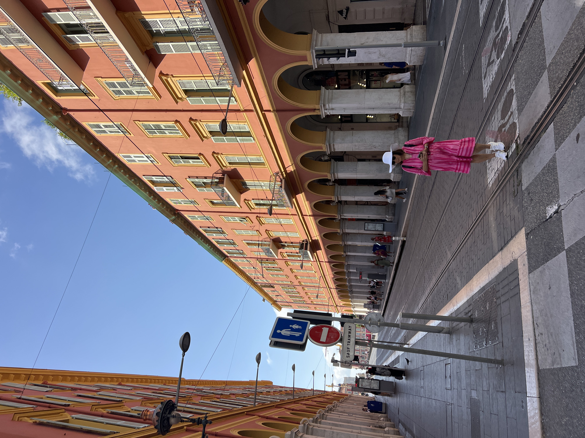 A wide shot captures a woman in a bright pink dress and a white sun hat walking across tram tracks on a street in Nice, France. She is holding a phone in her right hand and looking down. The street is lined with buildings painted in shades of coral and yellow, featuring arched walkways and balconies. A tram passes by in the distance. The sky is clear and blue with scattered clouds. Several traffic signs are visible, including a pedestrian crossing sign and a "Sauf Tramway" (Except Tramway) sign. The pavement is made of gray cobblestones, and there are tram tracks running down the center of the street. People can be seen walking on the sidewalks and near tram stops. The overall scene depicts a sunny day in a European city.