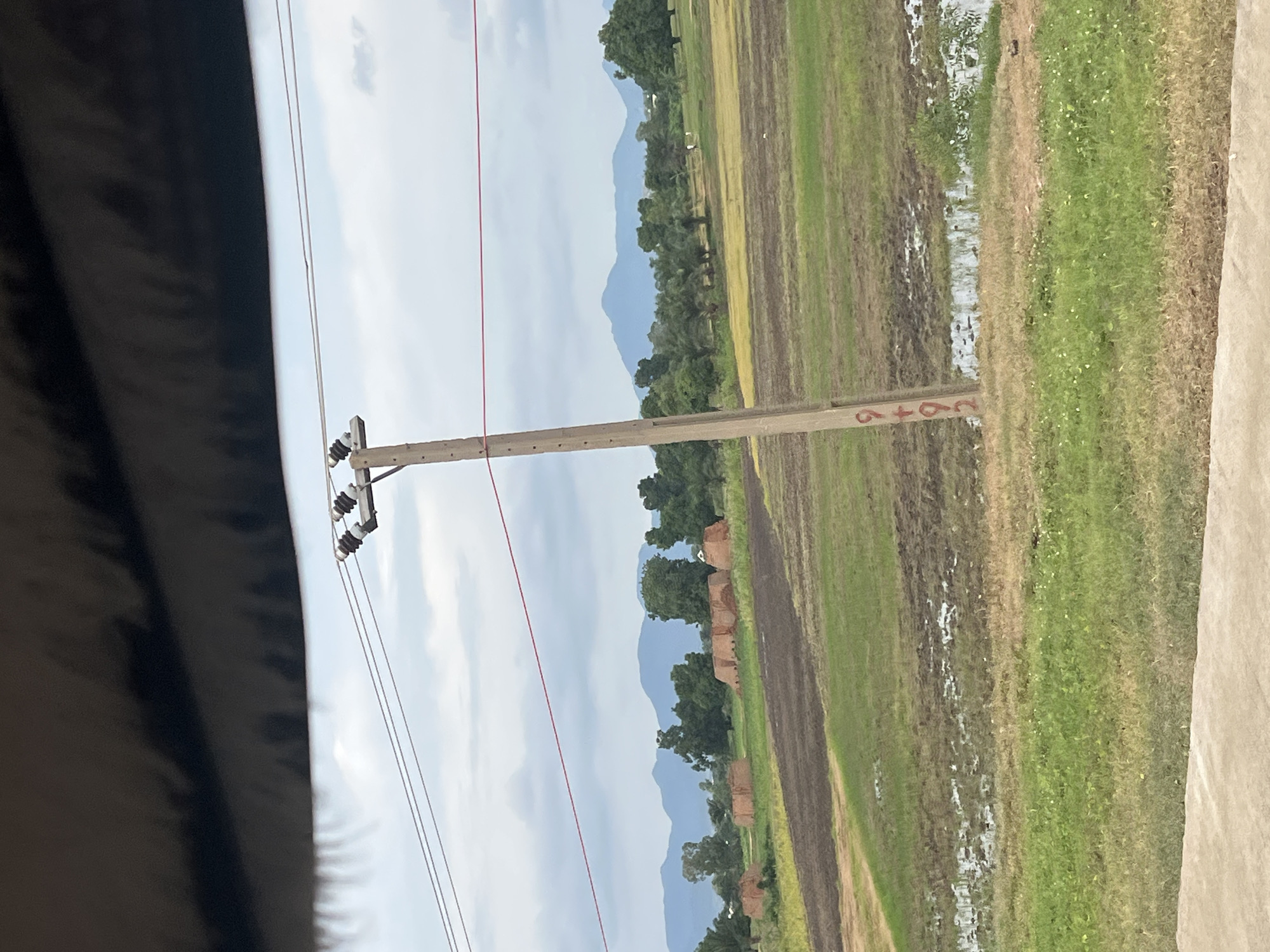 The image depicts a rural landscape with a utility pole in the foreground, likely located in Jalingo, Nigeria. The pole is wooden and has red markings, possibly a number or code, indicating "9+92". Wires are strung from insulators attached to the top of the pole, extending across the frame. In the middle ground, a field is visible with rows of what appear to be unharvested crops or tilled soil, interspersed with patches of water. Further in the distance, several brick structures, possibly kilns or temporary dwellings, are scattered across the landscape. Beyond the fields, lush green trees and vegetation are present, with rolling hills and mountains forming the backdrop under a cloudy sky. The overall scene suggests an agricultural setting.