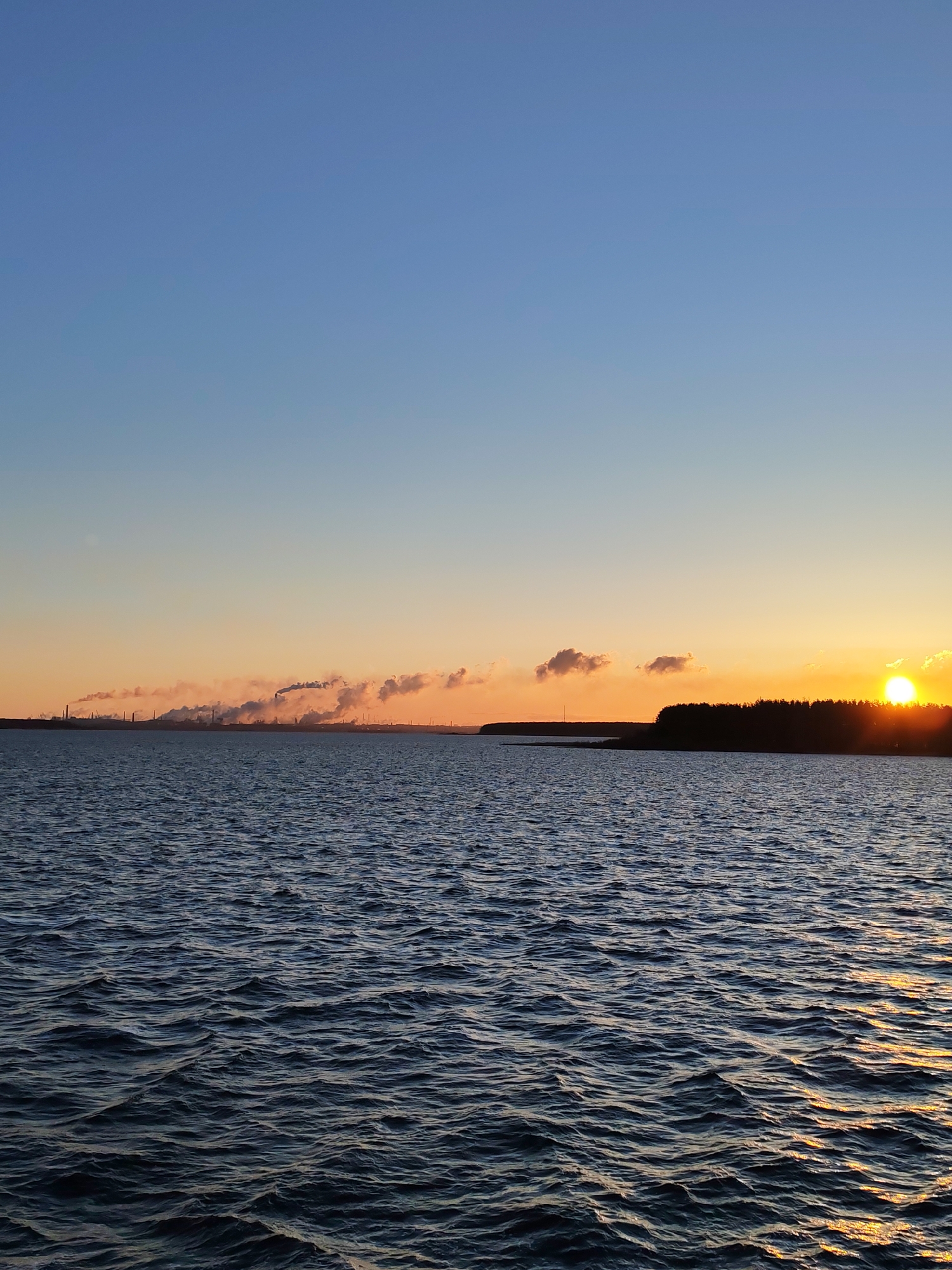 The image depicts a body of water with gentle waves, possibly a lake or sea, under a clear sky transitioning from blue to orange as the sun sets. Silhouettes of what appear to be industrial structures, some emitting smoke, are visible on the horizon to the left. To the right, a darker mass possibly representing a shoreline or forested area obscures the setting sun. The location in the city is unknown.