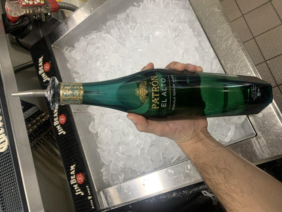 A person holds a bottle of Patrón El Alto tequila, green with gold lettering, with a spout attached. The bottle is positioned horizontally over a stainless steel ice bin filled with crushed ice. In the background, black Jim Beam bar mats are visible next to a metal dispenser with red accents. The scene appears to be a bar setting.