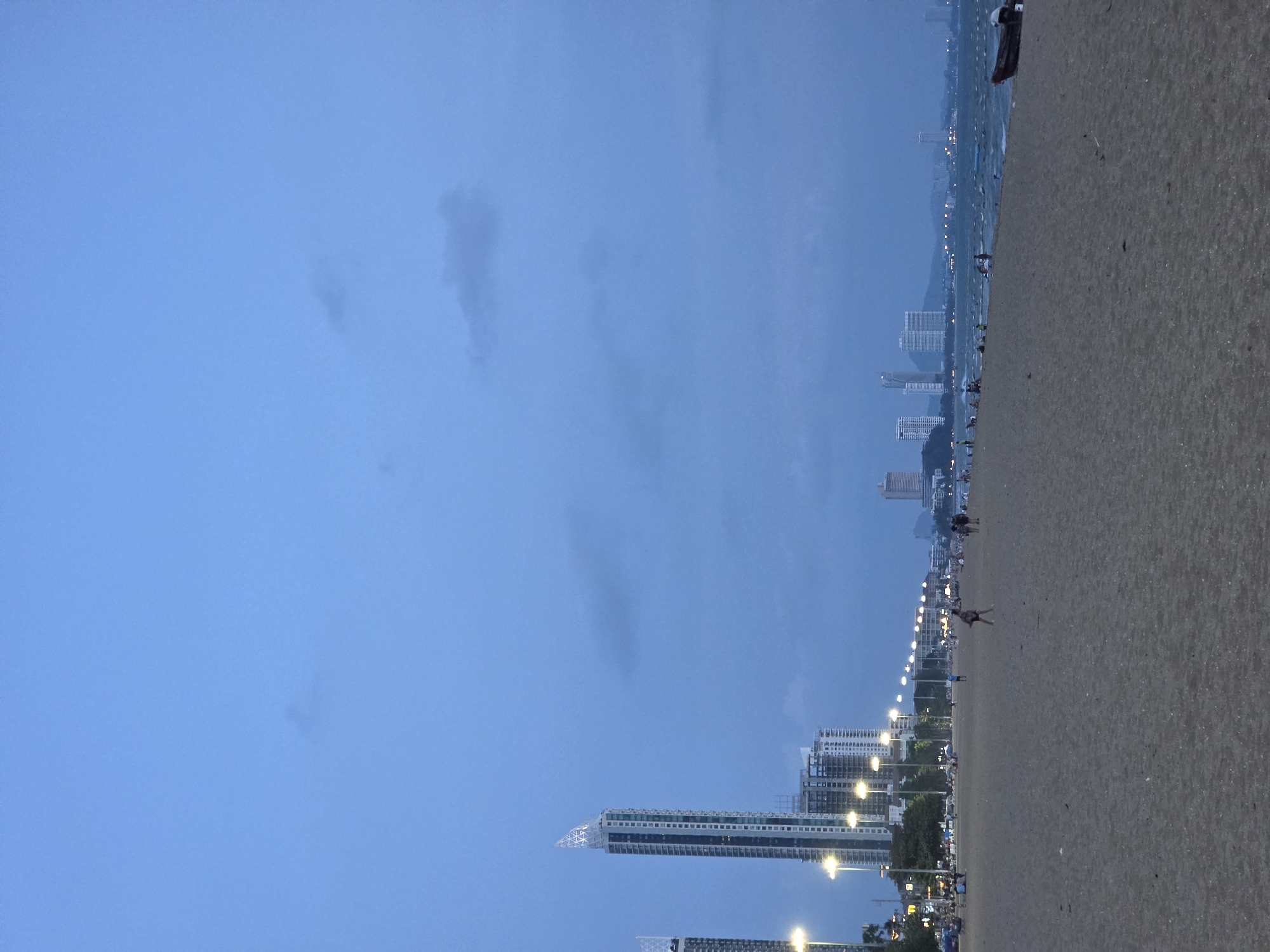The image portrays a wide urban beach scene at dusk in Pattaya City, Thailand. The dominant features include a vast, deep blue sky with faint cloud formations, a sprawling sandy beach, and a city skyline illuminated by evening lights.
The sky occupies a significant portion of the upper frame, displaying varying shades of blue characteristic of twilight, with subtle, wispy dark clouds. Below the sky, running horizontally across the scene, is a wide, light-brown sandy beach that stretches into the distance. A thin strip of the ocean is visible between the beach and the distant city, reflecting the dim light.
Along the coastline, numerous high-rise buildings and skyscrapers form a dense city backdrop. Many of these buildings show illuminated windows, indicating the evening hour. A prominent row of streetlights lines the edge of the beach, casting bright circles of light onto the sand and extending far down the shoreline, defining the path.
Several people are scattered along the expansive beach, appearing as small figures due to the distance. They seem to be leisurely strolling or relaxing, enjoying the evening ambiance. One figure in the mid-ground appears to be lying down or stretching on the sand. A dark, elongated object, possibly a small boat or wooden structure, rests on the sand near the water’s edge in the upper right portion of the beach.
The scene captures a peaceful and serene evening atmosphere at an urban beach. The low light conditions are evident from the dark blue sky and the dependence on artificial illumination from the streetlights and buildings. No text is visible in the image.