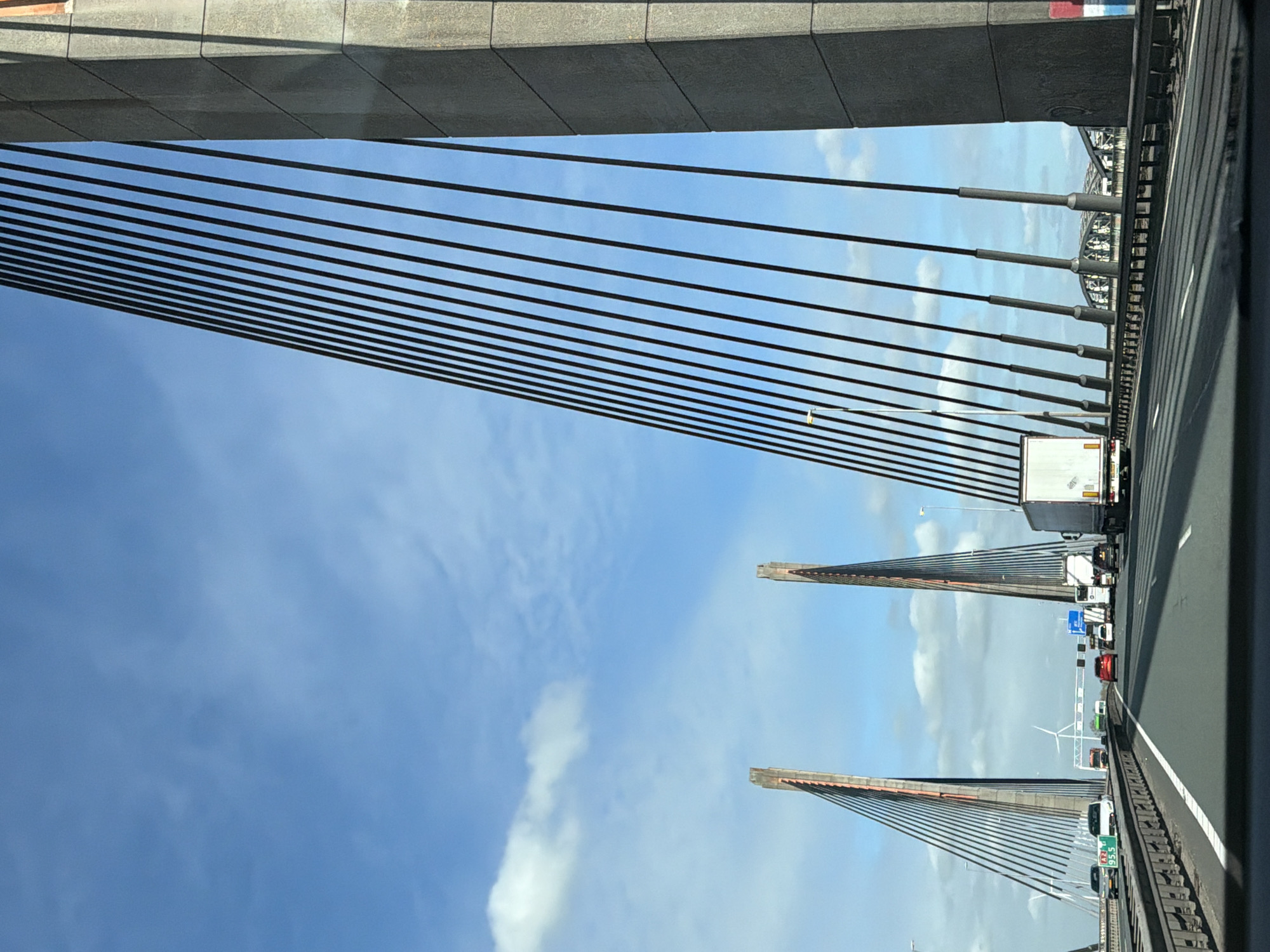 The image shows a view from a vehicle traveling along a cable-stayed bridge, likely the Martinus Nijhoff Bridge over the Waal River in Zaltbommel, Netherlands. The bridge's suspension cables are visible, anchored to two towers in the distance. A highway runs along the bridge, filled with moving vehicles, including trucks and passenger cars. The sky appears blue with scattered clouds. Overhead, the bridge's infrastructure is visible.