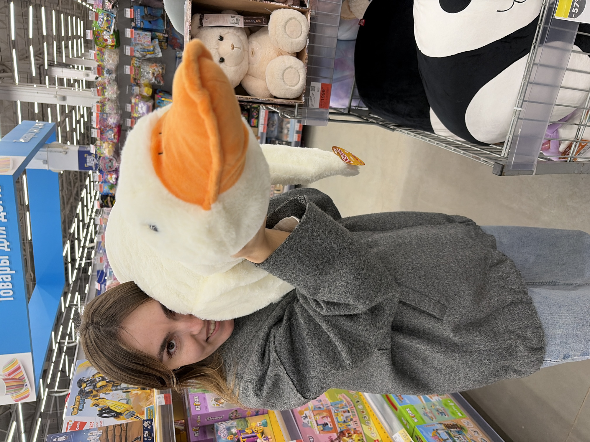 A woman in Izhevsk, Russia is shown in a retail environment, holding a large plush goose with an orange beak and a white body close to her face. She is wearing a gray jacket and light blue denim jeans.
She is positioned near shelves filled with toys and games. One shelf is visible with Lego sets and other toys. Other plush toys, including a white bear and a black and white yin-yang symbol are displayed in a wire basket above the toy shelf.
Retail signs in Russian are visible, suggesting the locale. The lighting overhead is bright and fluorescent.