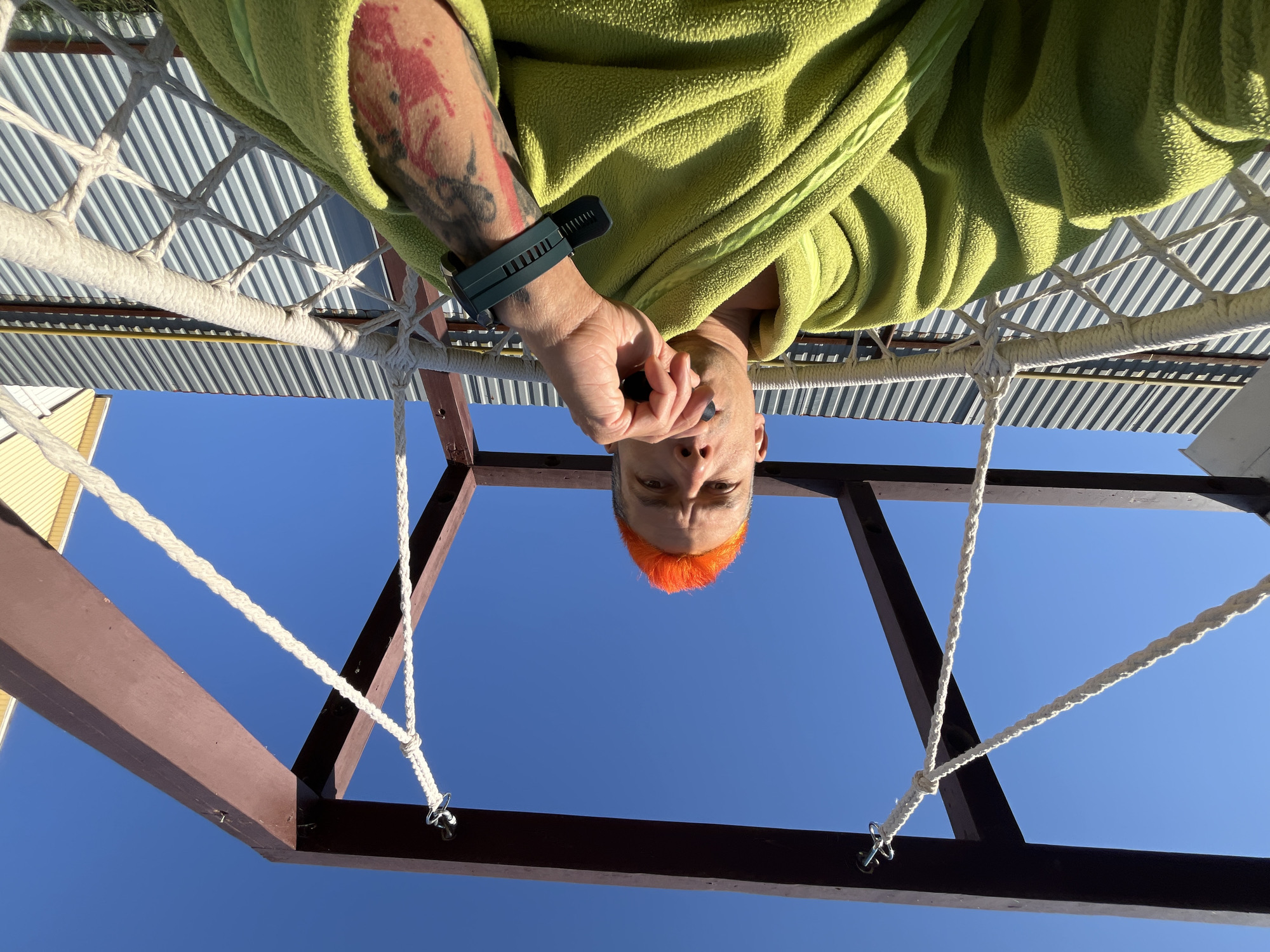 A low-angle, upside-down shot shows a person with orange hair looking down at the camera. The person is wearing a green fuzzy robe and a smartwatch on their left wrist, which has a tattoo. They are holding something small and dark in their hand. The person is positioned within a brown metal frame suspended by white ropes. Above the frame is a white-colored slatted roof. The background is a clear blue sky. The location is Chaikovsky, Russia.