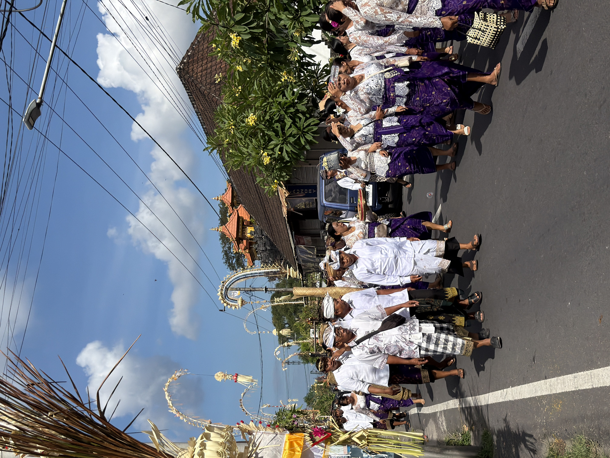 In Mengwi, Indonesia, a procession moves along a paved road under a bright blue sky with scattered clouds. The participants, dressed in traditional attire, are walking from right to left. Men in the front wear white shirts and patterned sarongs, some with headscarves. Behind them, women are adorned in matching purple sarongs and white lace tops. Several women in the foreground are holding their hands to their foreheads, shielding their eyes from the sun.
Decorative arches, possibly made of palm fronds, line the left side of the road, some adorned with white and yellow decorations. In the background, traditional Indonesian architecture with tiled roofs is visible. A blue van is parked on the right side of the road, and electrical wires crisscross the sky. The overall scene suggests a cultural or religious event taking place in a residential or urban area.