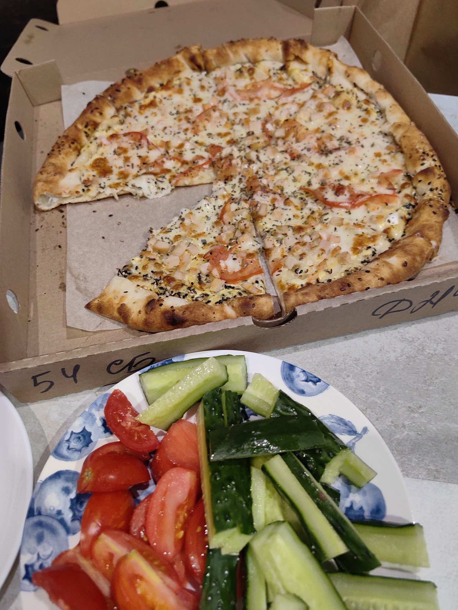 An overhead, medium shot captures a pizza in a cardboard box and a plate of chopped cucumbers and tomatoes. The pizza is a large, circular pie that has been sliced into multiple pieces and is topped with melted cheese, sliced tomatoes, and small pieces of what appears to be chicken or shrimp. The crust is golden brown and appears to be sprinkled with sesame seeds. The pizza box is open, revealing the pizza inside. To the right of the pizza, a plate of chopped cucumbers and tomatoes is visible. The vegetables are fresh and appear to be ready to eat. The scene is set on a light-colored, textured surface that could be a table or countertop. The overall impression is that of a casual meal or gathering.