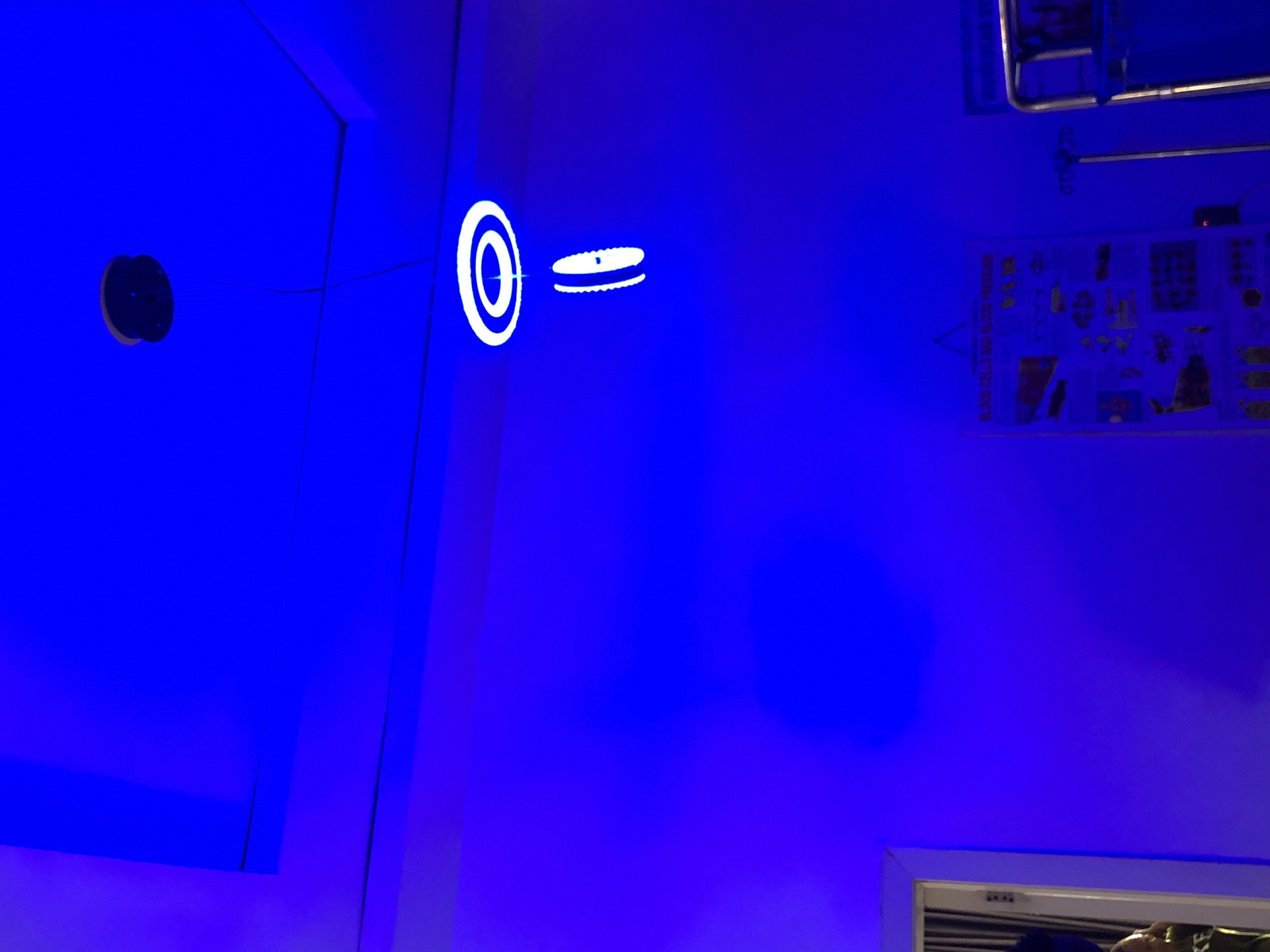 The image captures an indoor scene bathed in a deep, artificial blue light, illuminating walls and a ceiling. No people are visible.
On the left side, a black circular fixture protrudes from the blue surface, with thin wires extending horizontally from it across the wall. Towards the center, two distinct white, glowing circular objects are prominent. The object on the left is a hollow ring, while the one on the right is a flat disk with a glowing perimeter and a bright central dot, appearing suspended in the blue space.
In the upper right portion of the image, a metallic object, possibly part of a railing or specialized equipment, is partially visible, attached near the ceiling. Below it, a poster or chart is affixed to the wall. Though dimly lit by the blue hue, the title "BLOOD CELLS AND BLOOD PRESSURE" is discernible on this poster, suggesting a scientific, medical, or educational setting, such as a laboratory, clinic, or a science museum exhibit.
In the bottom right corner, a white-framed opening, possibly a window or a vent with horizontal slats, is seen. The overall environment, with its strong blue illumination and specialized objects, creates a futuristic or highly specialized atmosphere. There is no activity taking place; the scene shows static installations.