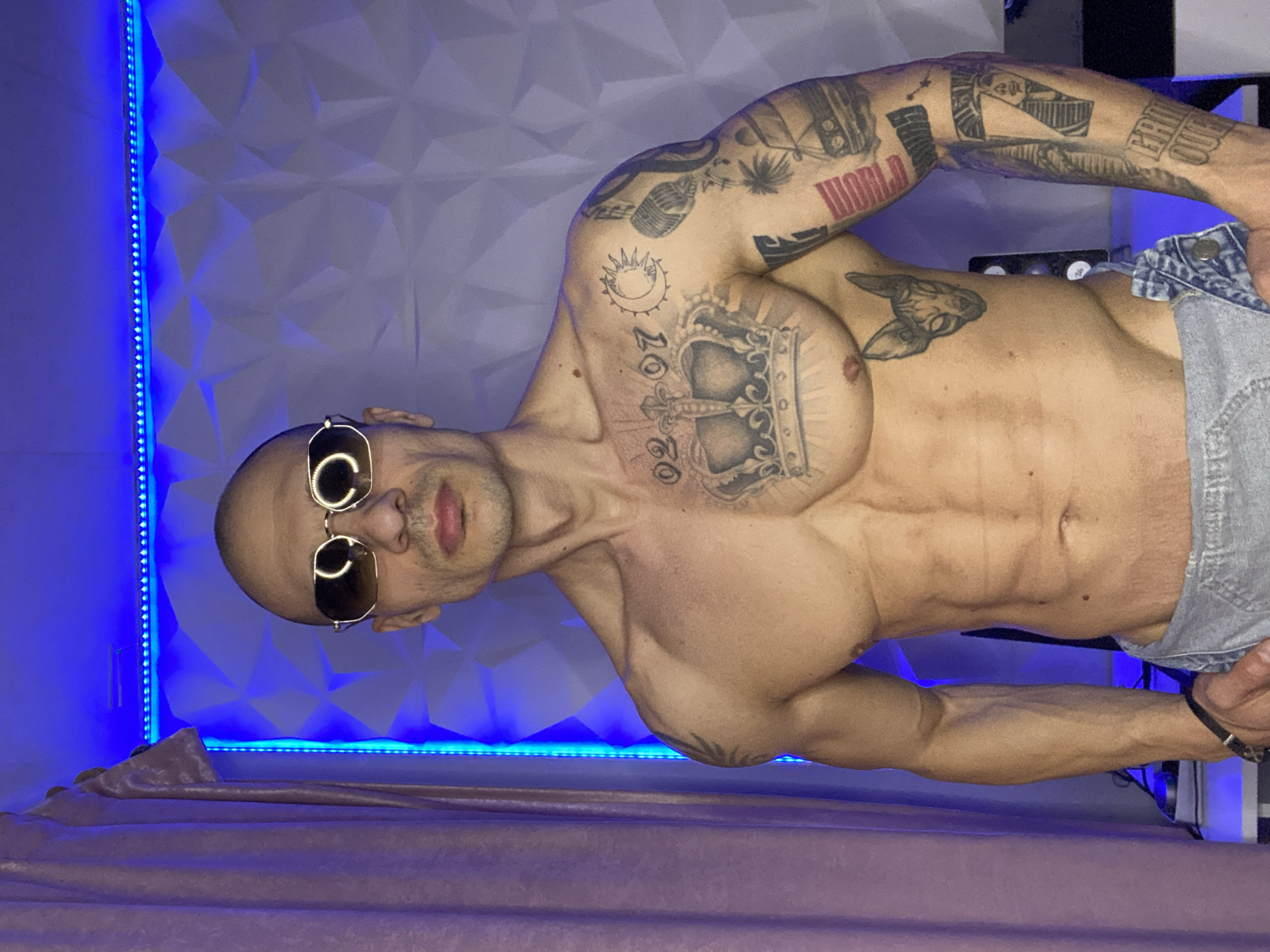 An adult male with a bald head and muscular physique stands shirtless, facing the camera. He wears mirrored aviator-style sunglasses and light-wash denim pants, unfastened at the waist. Tattoos are prominent across his upper body: a large crown design with the numerals "02 07" on his chest, a sun and crescent moon on his left shoulder, a Sphynx cat head on his left abdomen, and multiple designs covering his left arm, including a microphone, a car, a palm tree, and partially visible text such as "WORLD" and "GRIN." A faint tattoo is also present on his right shoulder. The background consists of a wall paneled with white geometric 3D shapes, illuminated by horizontal and vertical blue LED strip lighting. A light pink curtain hangs on the far left. Obscured dark objects are visible in the lower left foreground. The precise location within Pleven, Bulgaria, is not identifiable from the image.